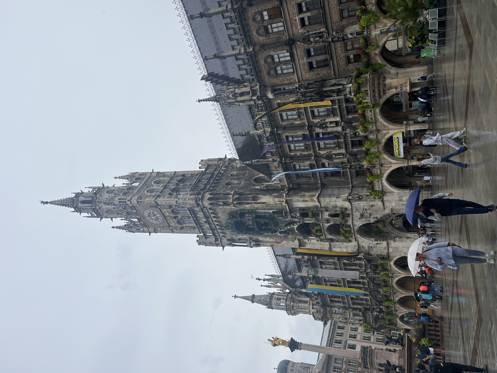 The image captures a wide-angle view of the Marienplatz in Munich, Germany, on a cloudy and rainy day. In the center of the frame stands the New Town Hall (Neues Rathaus), a magnificent Gothic Revival building with an ornate facade and a prominent clock tower. The tower features a historical Glockenspiel, which is currently not in motion. Several flags, including the Bavarian flag, are displayed on the building. To the left of the Town Hall, the Mariensäule (Mary's Column) is visible, topped with a golden statue of the Virgin Mary. In the background, the dome of St. Peter's Church can be seen.
The square in front of the Town Hall is wet and reflective due to the rain, with several people walking around, some holding umbrellas. A woman in the foreground is holding a white umbrella and wearing a light blue top and light-colored pants. To her right, a man is holding a dark blue umbrella and wearing a dark vest over a blue shirt and dark pants. Other individuals are seen in the middle ground, engaged in various activities such as walking and taking pictures. The overall atmosphere is one of a typical European city square, despite the inclement weather.