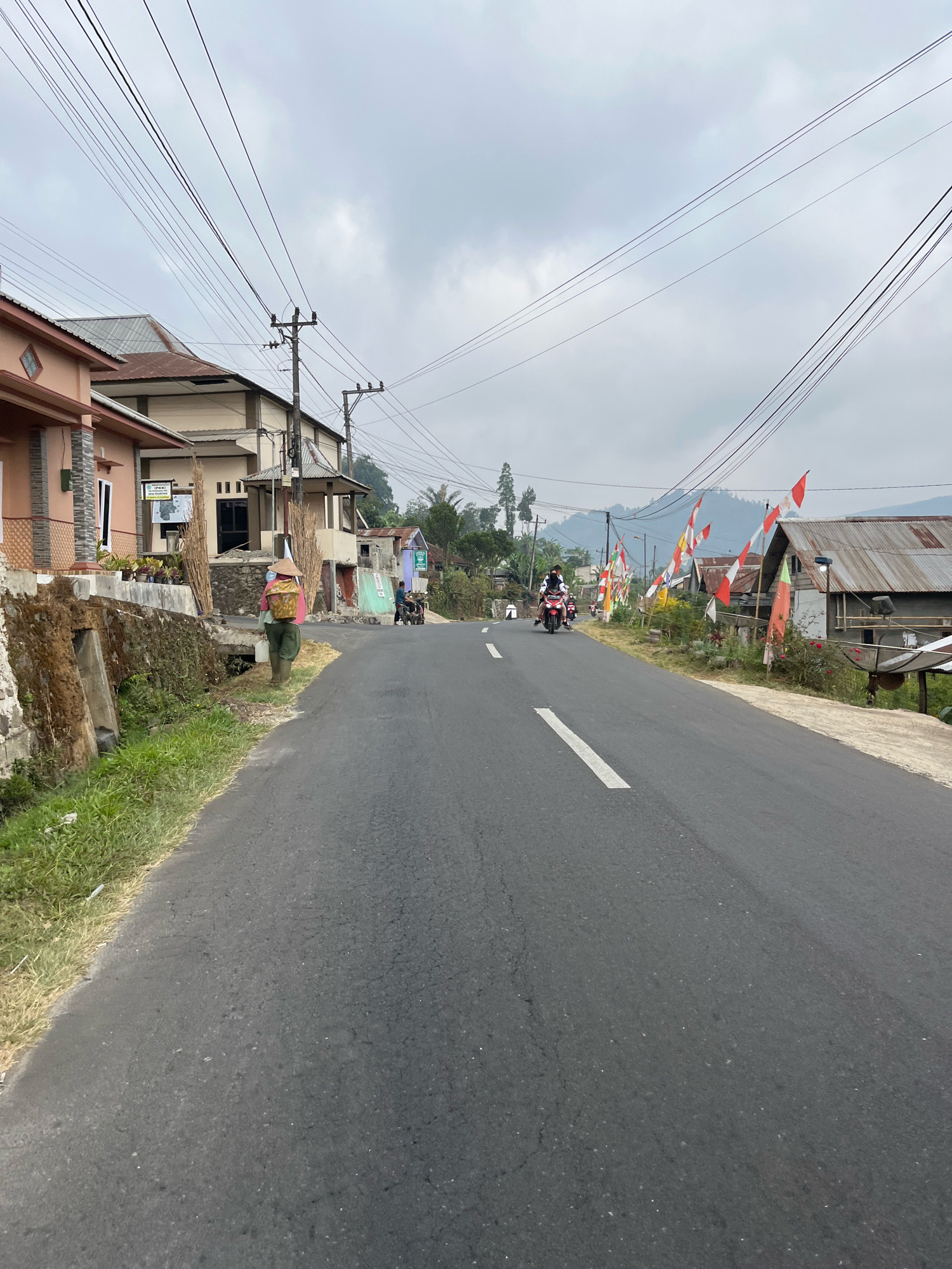The image depicts a paved road in a rural or suburban area, likely in Balun, Indonesia, given the context. The sky is overcast with clouds. On the left side of the road, there are several houses with varying architectural styles. Some have pink and beige facades, while others appear to have corrugated metal roofs. A person wearing a wide-brimmed hat and green pants is walking on the left side of the road, carrying a basket on their back and a white bucket in their hand. They are walking away from the camera, towards the direction of the road.
The road itself is asphalt and has a white dashed line marking the center. Several motorcycles are visible on the road, with riders. On the right side of the road, there are more structures, including some with metal roofs and what appears to be a satellite dish. Numerous Indonesian flags, predominantly red and white, are displayed along the right side of the road, suggesting a festive occasion or national commemoration. In the background, rolling hills and trees are visible, receding into the hazy distance. Utility poles with numerous wires run along the road. The overall impression is a scene of daily life in a community, possibly during a period of celebration.