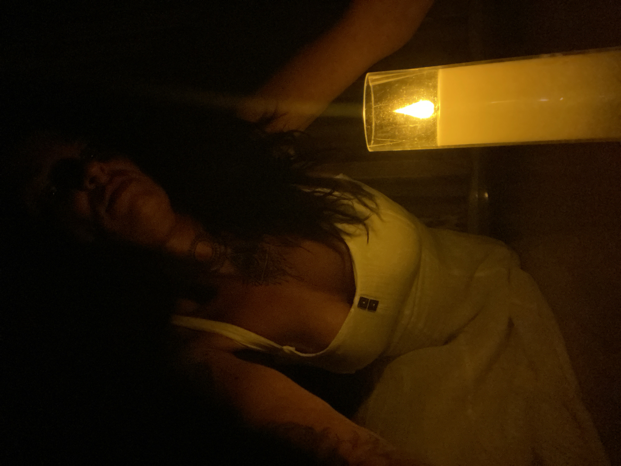 A person is illuminated by a single candle in a dark environment. The person is wearing a white dress with straps and a low neckline. The dress appears to have two square buttons on the front. Their arm is visible on the left side of the frame, and their face is partially obscured by shadow, looking upwards. A tall, clear glass candle holder containing a lit candle is positioned in the foreground on the right side of the image. The candle flame is bright and casts a warm glow on the scene. The background is entirely dark, suggesting an indoor or nighttime setting. There are no visible elements that can be used to determine the location in Mũi Né, Vietnam.