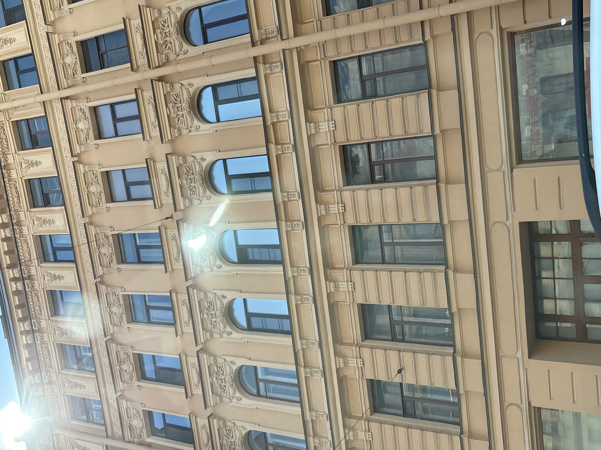 The image displays a close-up, low-angle view of the facade of a historic building in Saint Petersburg, Russia, with a bright sun creating lens flare and reflections on some windows. The building exhibits ornate architectural details, including decorative moldings, cornices, and pilasters surrounding numerous windows. The windows are of various shapes and sizes, with some being rectangular and others arched, all featuring dark frames. The facade is painted a pale yellow or beige color. Several floors of the building are visible, each with multiple windows. The sky, visible at the top of the image, is bright and sunny with some light clouds. The architecture suggests a building from the 19th or early 20th century, typical of St. Petersburg's historic city center.