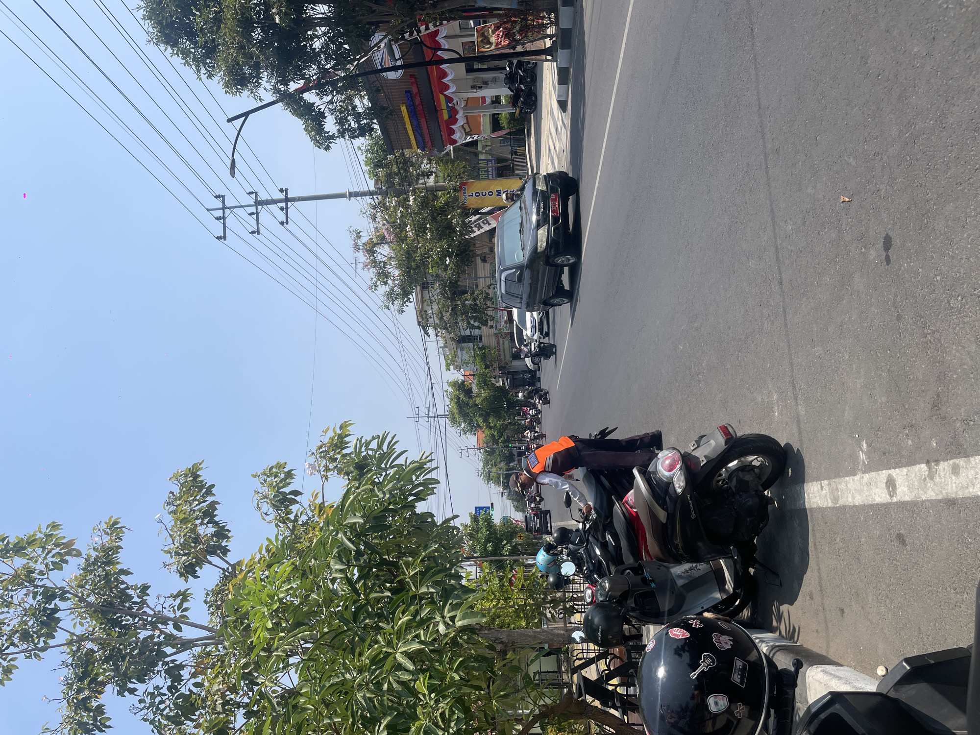 The image captures a street scene in Bojonegoro, Indonesia, on a clear, sunny day. In the foreground, several parked motorcycles, one black with stickers and another with a blue helmet, are positioned along a sidewalk. A man wearing an orange vest and a cap stands next to a motorcycle, appearing to be interacting with it or its owner. He is dressed in dark pants and a light-colored shirt.
The street itself is paved with asphalt and features a white line marking a lane. Traffic is visible, including a dark green SUV with a license plate (S 2223 MA) on the right side of the road, and several other motorcycles and cars in the distance, suggesting moderate traffic flow.
Lining the street are trees with lush green foliage, some with small pink blossoms. Utility poles with numerous electrical wires are prominent against the bright blue sky. Buildings with awnings and signage are visible on the right side of the road. The overall environment suggests a typical urban street in Indonesia, with a mix of parked vehicles, moving traffic, and pedestrian infrastructure.