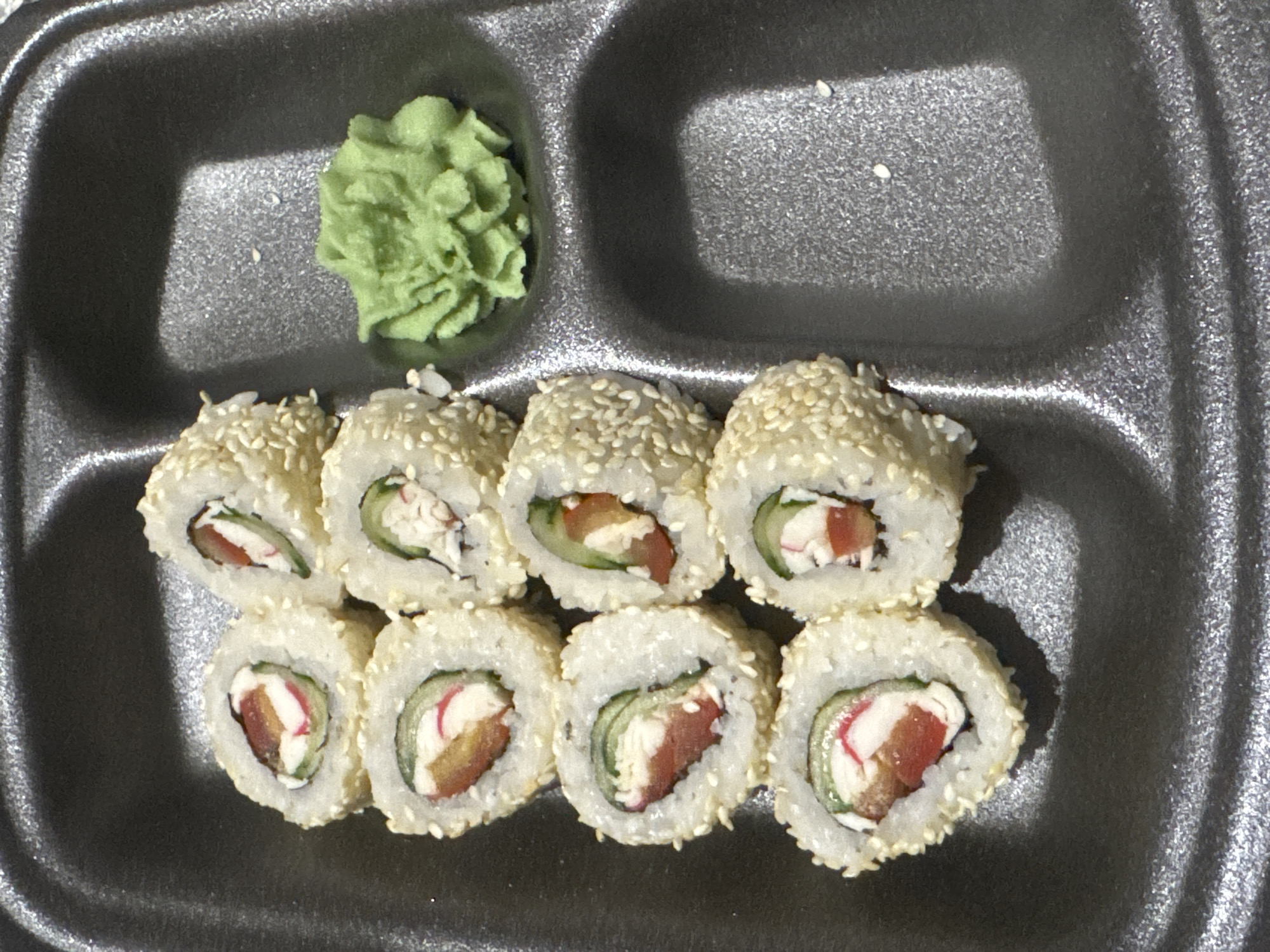 A black styrofoam container holds eight pieces of sushi rolls covered in sesame seeds and a dollop of bright green wasabi paste. The sushi rolls are arranged in two columns of four, with the wasabi paste positioned to the right of the rolls in a separate compartment of the container. The rolls appear to contain ingredients such as cucumber, red pepper, and crab stick. The image was taken indoors, likely in a residential setting in Zaokskiy, Russia, as suggested by the context.