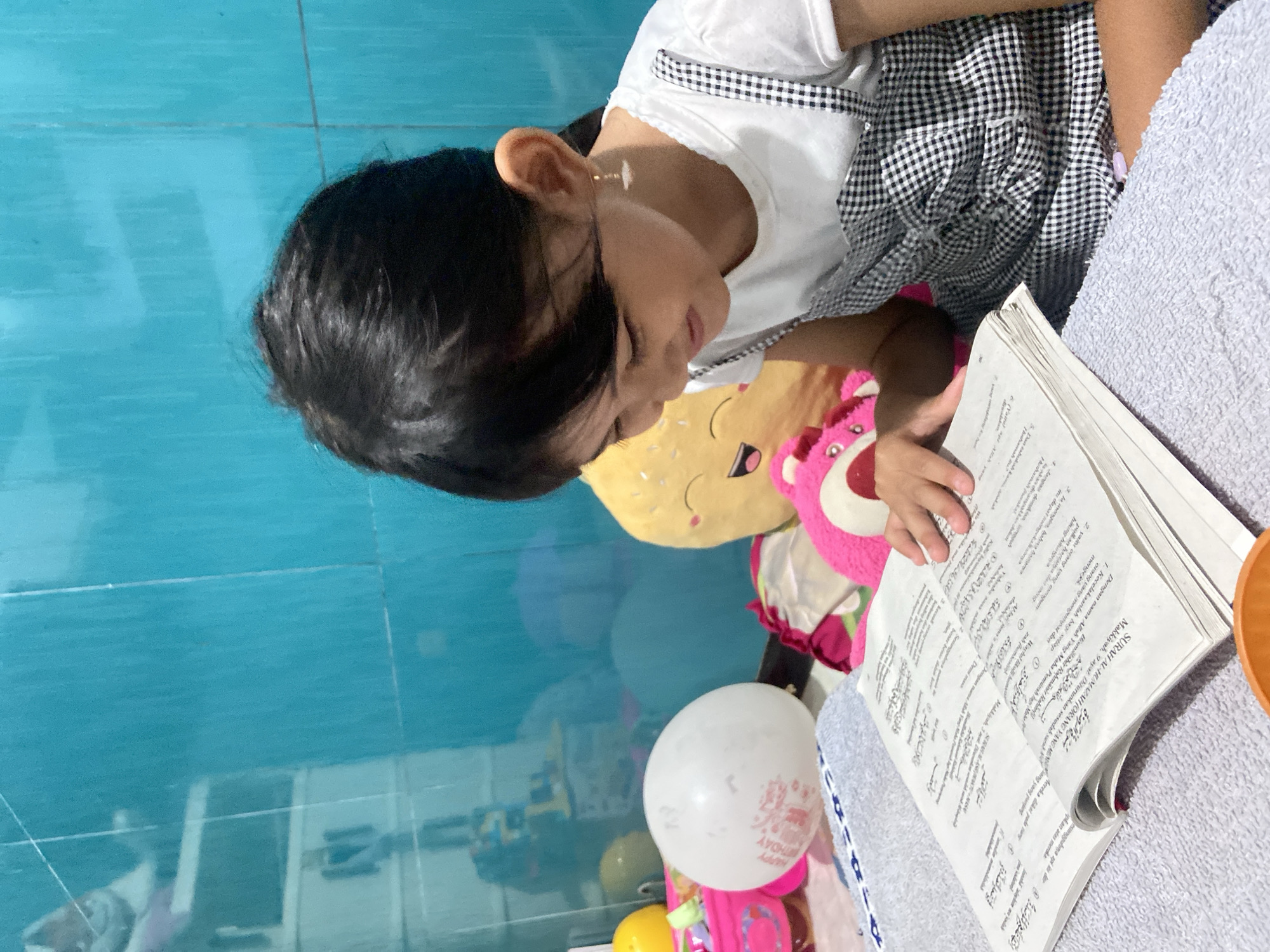 A young girl is reading a book written in Arabic in a room with turquoise tile flooring. The girl has dark hair and is wearing a white shirt with a black and white checkered dress. She has a yellow plushie with a smiling face and a pink bear next to her. There is a white balloon that says "HAPPY BIRTHDAY" in pink letters. The girl appears to be studying or reading the book intently. The location is likely Gelam, Indonesia, based on the information provided.