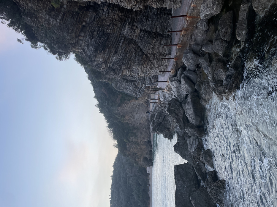 The image depicts a coastal scene along a rocky shoreline in Budva, Montenegro, specifically on the path leading towards Mogren Beach. The environment is characterized by a dark, turbulent sea on the left, breaking against numerous large, grey boulders that form the immediate coastline. A narrow, paved concrete path with several rusty metal posts embedded along its edge runs parallel to the shore, curving from the mid-ground towards the background, directly beneath steep, layered dark grey cliffs. These cliffs, occupying the right side and background, are heavily stratified horizontally and topped with dense green vegetation, including trees and bushes. The sky overhead is a pale, hazy blue, transitioning to a lighter shade near the horizon.
Two individuals are visible on the path. One person, dressed in dark clothing, is in the mid-distance, walking away from the viewer along the path. A second, more distant figure is positioned closer to a small, light-colored sandy beach area visible further along the coast, with what appears to be a small structure at its edge. The lighting suggests either early morning or late afternoon conditions.