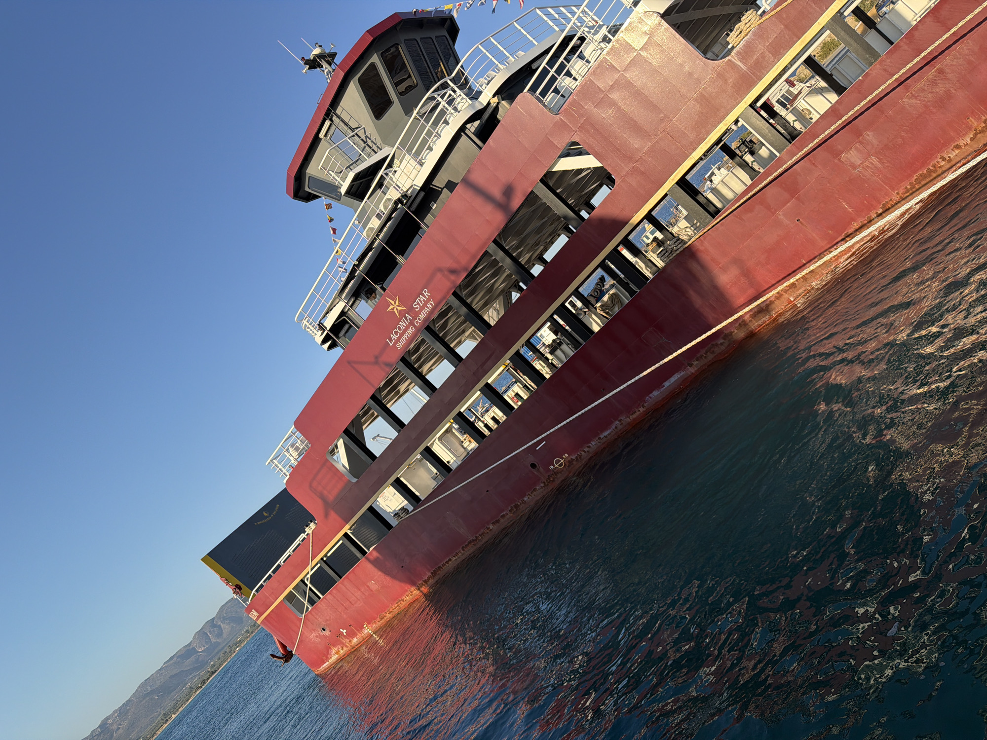 The image shows a tilted red ferry boat, the "Laconia Star," partially submerged in blue water. The ferry has multiple decks with windows. The boat appears to be listing heavily to the port side. The scene is set against a clear blue sky and a backdrop of hills and coastline, suggesting a location near land. The water is calm with reflections of the boat visible. The overall impression is of a vessel in distress or capsizing. It's a clear sunny day.
