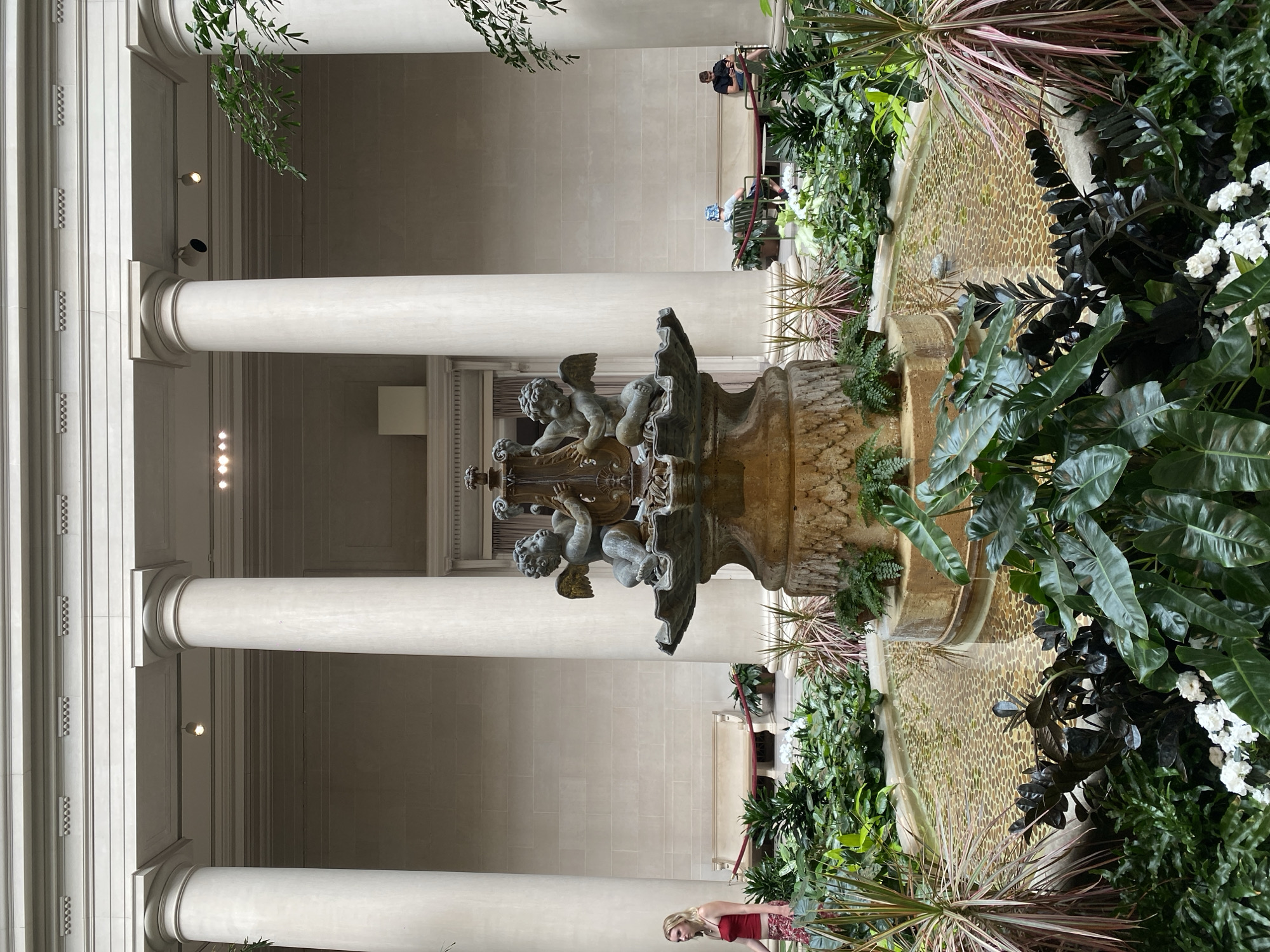 This photograph captures the interior courtyard of the National Gallery of Art in Washington, D.C. A classical-style fountain, adorned with two cherub sculptures, is the central focus of the scene. The fountain is surrounded by lush greenery and a shallow pool with a pebble-lined bottom. Tall, white columns support the building's architecture, creating a sense of grandeur and symmetry. Several visitors are present, with a woman in a red top walking past the fountain, and two individuals seated on a bench in the background. The lighting suggests it is daytime, possibly overcast or in a shaded area.