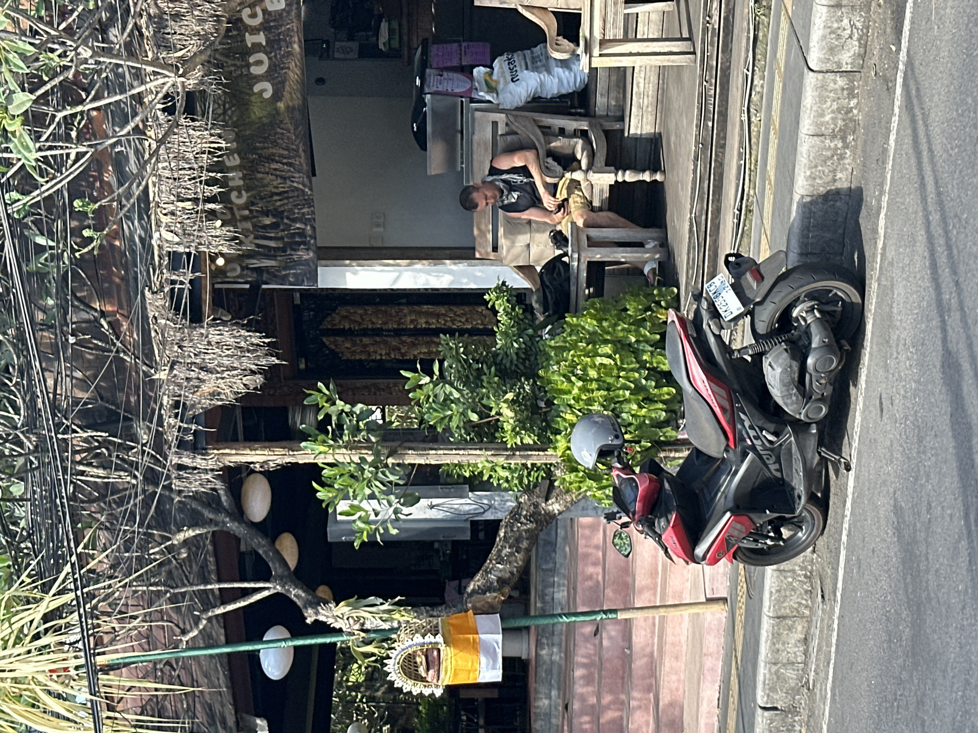 The image shows a street scene in the Ubud District, Indonesia. A red and black scooter is parked on the side of the road in front of a building with a thatched roof. A man is sitting on a wooden bench outside the building, looking at his phone. The building appears to be a restaurant or cafe, with a sign that says "KITCHEN JUICE BAR". There are plants and trees around the building, and a street lamp on the left side of the image. The scene is lit by bright sunlight, casting shadows on the ground. The scooter has a license plate that reads "DK 2558 KCB".