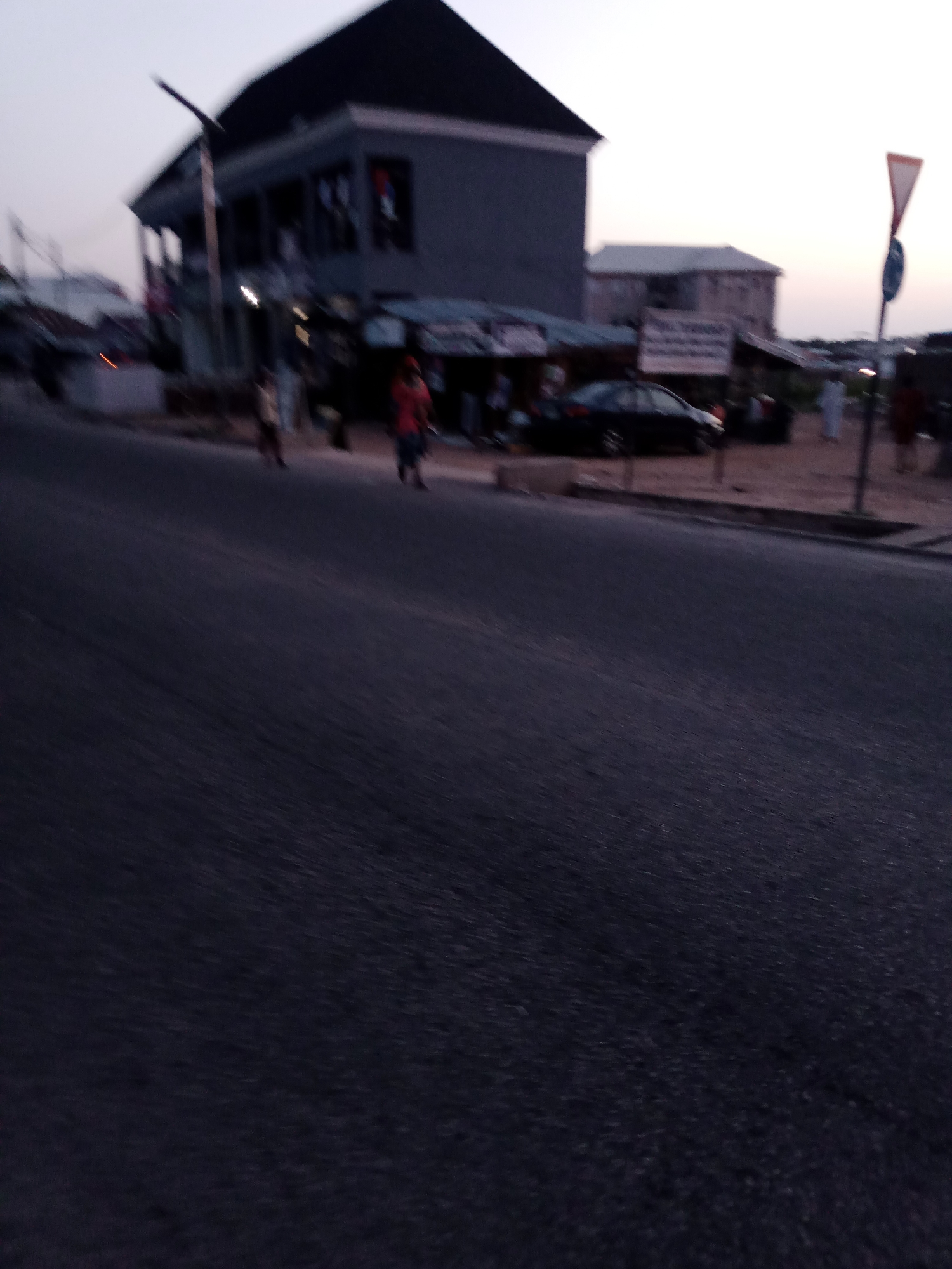 This is a street scene in Jalingo, Nigeria, at dusk. A dark asphalt road occupies the foreground, with a subtle gradient of shade and texture suggesting it's damp or reflecting ambient light. In the mid-ground, a two-story building with a dark pitched roof and grey walls stands prominently, appearing to be a commercial or residential structure. Windows are visible on its facade. To the right of this building, a smaller, lighter-colored building is situated. Several individuals are present on the sidewalk and road. One person in a red shirt is walking on the road, and others, appearing as blurred figures, are near the buildings and on the sidewalk. A dark-colored sedan is parked on the road in front of one of the buildings. Traffic signage is visible on the right side of the image, including a triangular warning sign. The sky is a pale, dusky grey, indicating the transition from day to night. The overall impression is one of a moderately busy street at the end of the day.