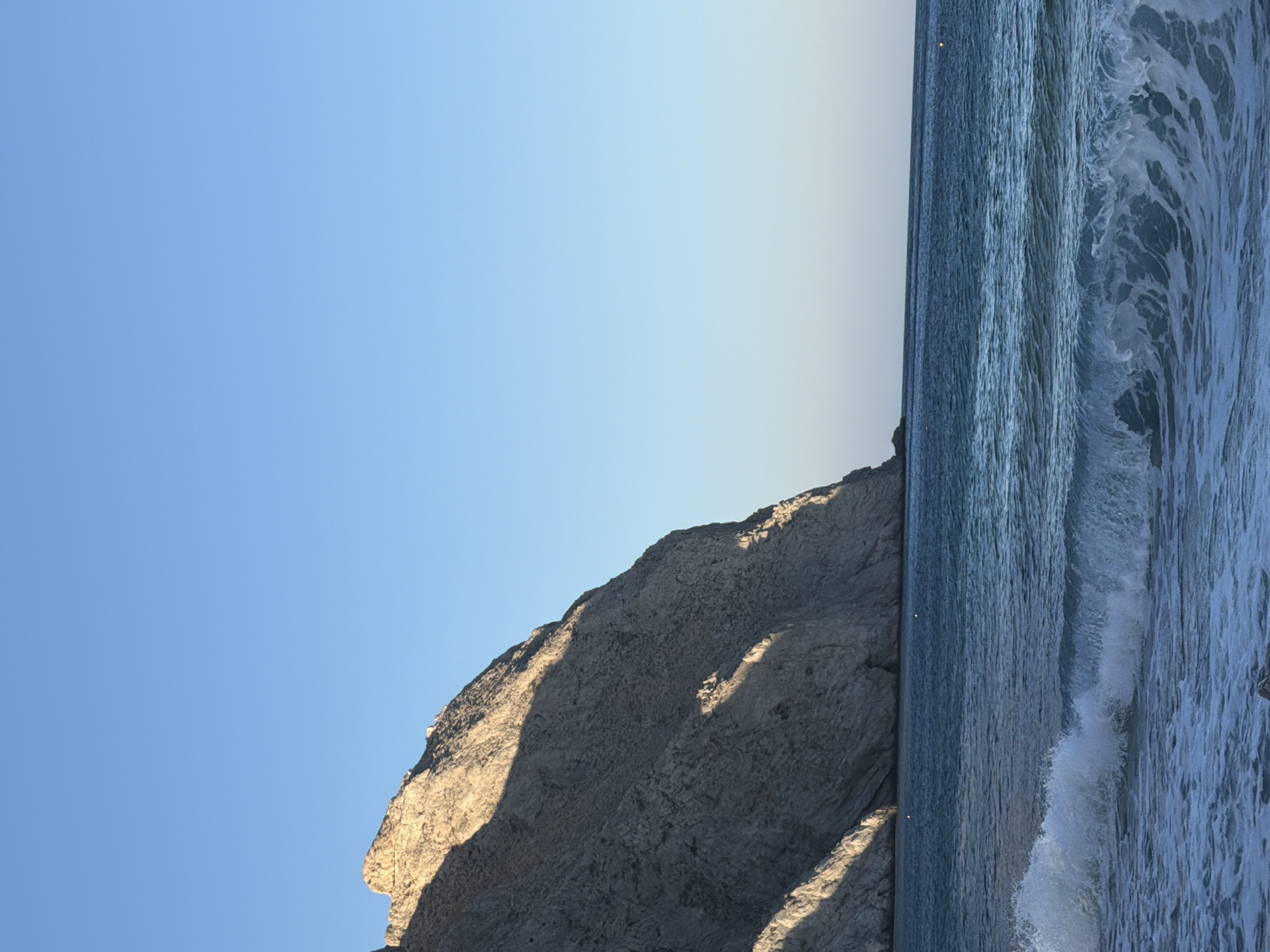 The image depicts a scenic coastal view with a large, rugged cliff face dominating the left side. The cliff is partially illuminated by sunlight, casting shadows across its textured surface. Below the cliff, the deep blue sea stretches towards the horizon, with gentle waves rolling towards the shore. A white foamy wave is breaking in the foreground, adding dynamism to the scene. The sky above is a clear, bright blue. The location is likely a beach on the island of Santorini, within the Thira Municipal Unit, Greece, given the characteristic volcanic rock formations and the Aegean Sea. There are no people or discernible man-made objects in the frame, besides what appears to be a small, distant boat or buoy in the water. The overall atmosphere is serene and natural.