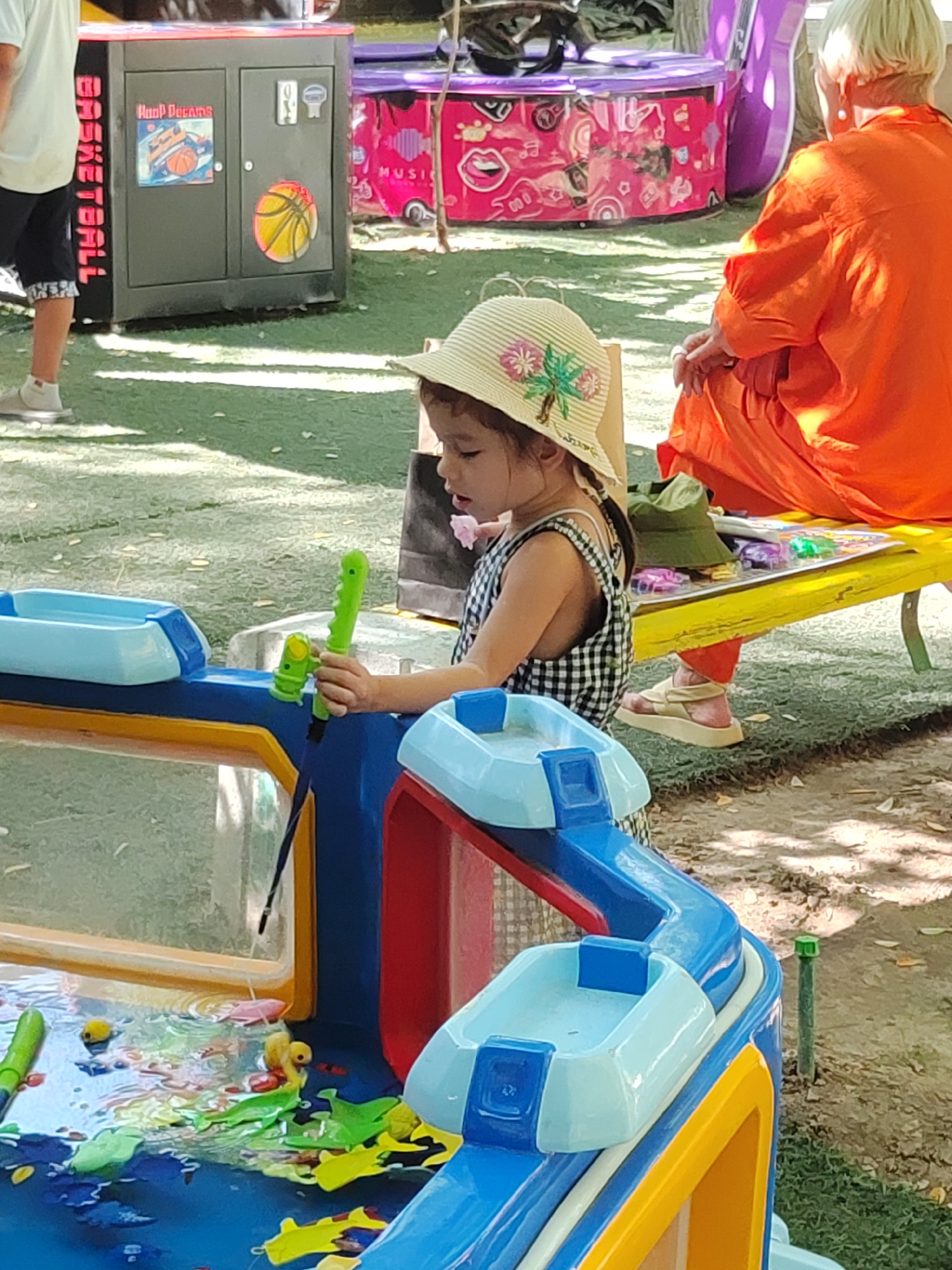 The image shows a young girl in a checkered dress and a straw hat playing with a green toy gun in a water table. She is focused on the water and toys in front of her. In the background, a person wearing an orange garment is sitting. Further back, there are colorful arcade machines. The ground is covered with green turf and scattered leaves, suggesting an outdoor setting, possibly a park or amusement area. The arcade machines have graphics that suggest a connection to basketball and music. The overall scene appears to be a recreational area, likely in an urban environment given the presence of arcade machines. Without specific landmarks, it is impossible to definitively place this scene in Tashkent, Uzbekistan, or any other specific city.