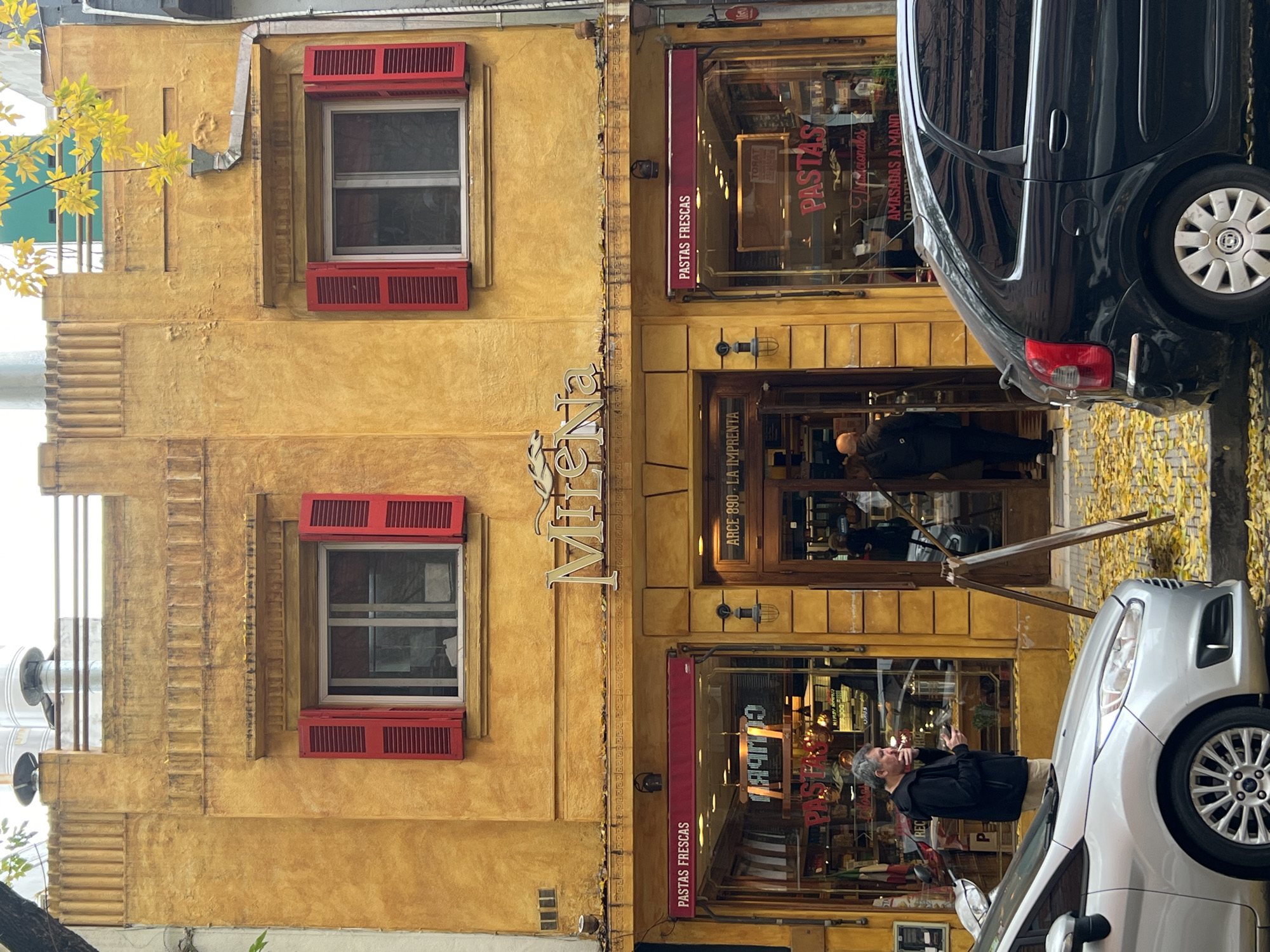Here's a description of the image:
The image shows the exterior of a building in Buenos Aires, Argentina, identified as a pasta shop called "Mireia Pastas Frescas," with the address "Arce 890 - La Imprenta." The building has a yellow facade with red window shutters. Two windows are visible on the second floor.
The shop's entrance is in the center of the frame, with glass doors and windows displaying pasta products and signage. People are visible inside and near the entrance.
On the right side of the image, partially obstructing the view of the shop, are parked cars. One is black, and the other is silver. A man stands near the silver car, appearing to smoke.
Fallen yellow leaves are visible on the street.
