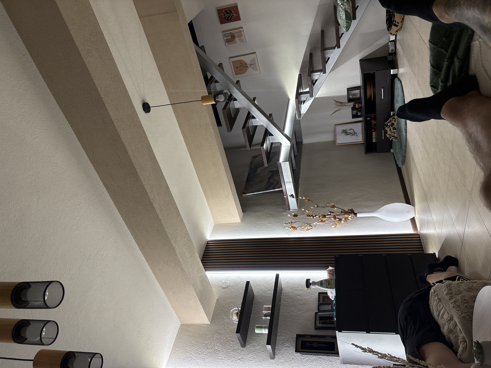 The image depicts an indoor scene, likely a living room, with a modern architectural design. A prominent feature is a sleek, multi-level staircase with dark treads and a metallic frame. The walls are textured, and the ceiling has exposed beams painted in a light brown and off-white color scheme. Decorative elements include framed artwork on the walls, a tall white vase with dried branches, and several shelves on the left side of the image displaying various objects.
On the left, a black chest of drawers is visible, topped with a few items, including a bottle. In the foreground, on the floor, two individuals are lying down, seen from the perspective of their legs and feet. One person is wearing black socks and is partially obscured by a textured cushion. The other person's legs are also visible, clad in black socks and resting on a dark green mat. The overall impression is of a relaxed, contemporary living space. The specific location in Asenovgrad, Bulgaria, is not visually inferable from the image content.