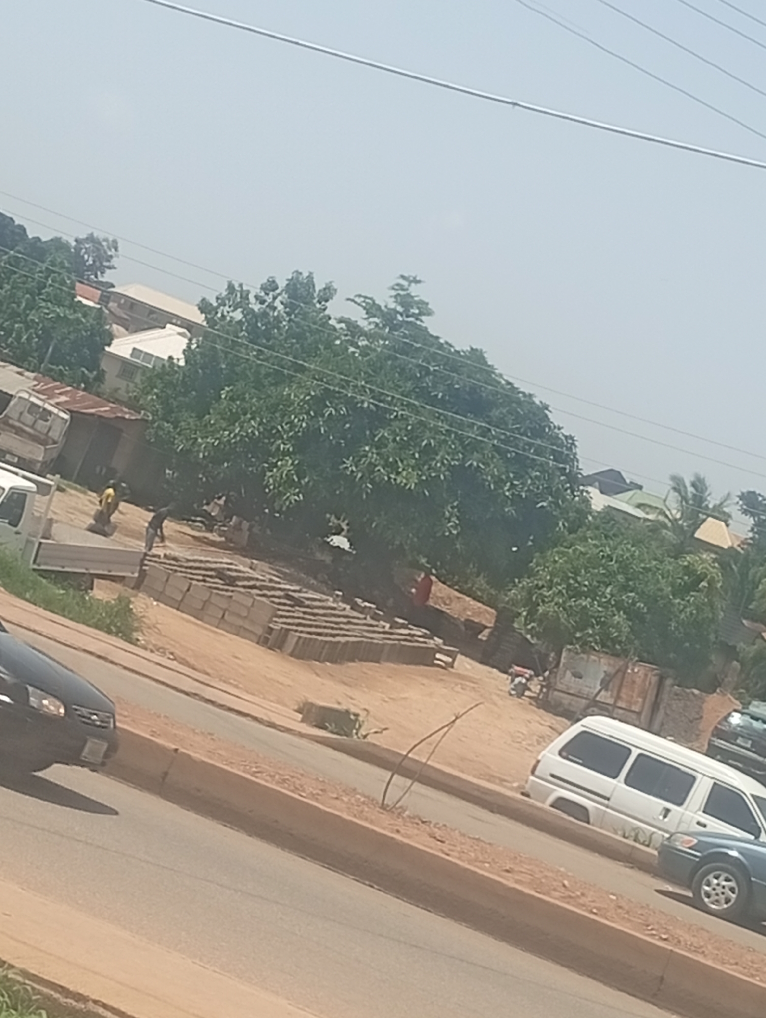 The media file depicts an outdoor scene in Bukuru, Nigeria, on a clear, sunny day. The foreground shows a paved road with a dirt shoulder. A dark-colored car is partially visible on the left, with its front bumper and headlights showing. Further along the road, a white van is parked, and next to it, a dark-colored sedan.
The background reveals a bustling area with several structures and a large, leafy tree dominating the mid-ground. To the left, a small building with a corrugated iron roof and a white truck with a flatbed are visible. In front of this building, two people are engaged in work. One person, wearing a yellow shirt, is bent over, possibly handling materials, while another person in a dark shirt is standing and appears to be interacting with them.
Adjacent to the people and the truck, a large quantity of grey bricks is neatly stacked, suggesting a construction or manufacturing site. The ground in this area is dusty and sandy. Beyond the tree, more buildings with varied roof colors, including green and yellow, are scattered amongst the lush vegetation. The presence of power lines stretching across the upper portion of the frame indicates an urban or semi-urban setting. The overall impression is one of everyday activity and commerce in a Nigerian community.