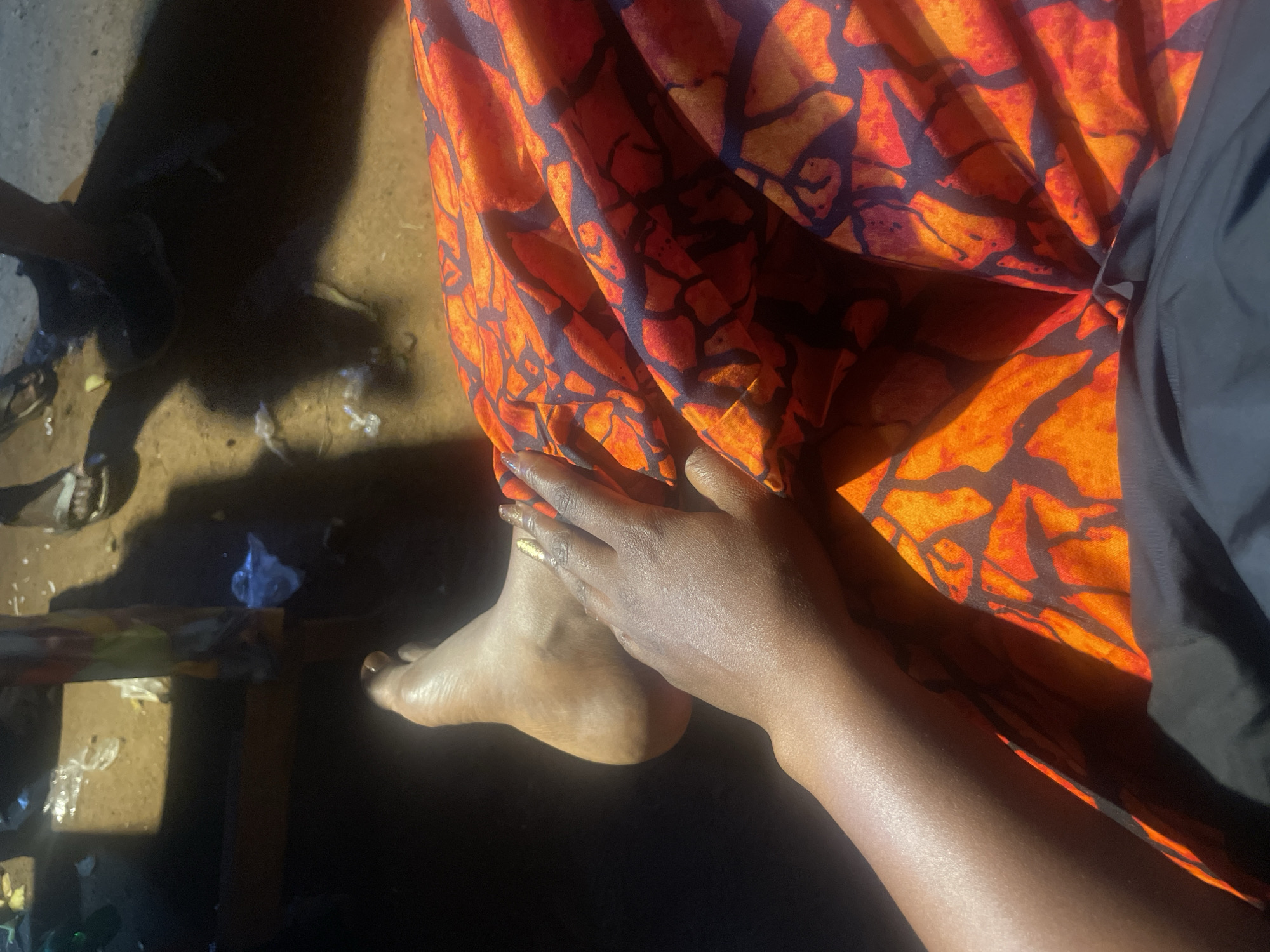 In the city of Du, Nigeria, a person's bare foot and lower leg are visible in the foreground, casting a shadow on the dusty, sun-drenched ground. They are wearing a vibrant orange and red patterned fabric, possibly a skirt or wrap. A hand, adorned with gold nail polish, rests on the fabric. The background is dimly lit and shows hints of discarded plastic debris and the legs of other individuals wearing sandals. The lighting suggests it is daytime, with strong sunlight creating sharp shadows.
