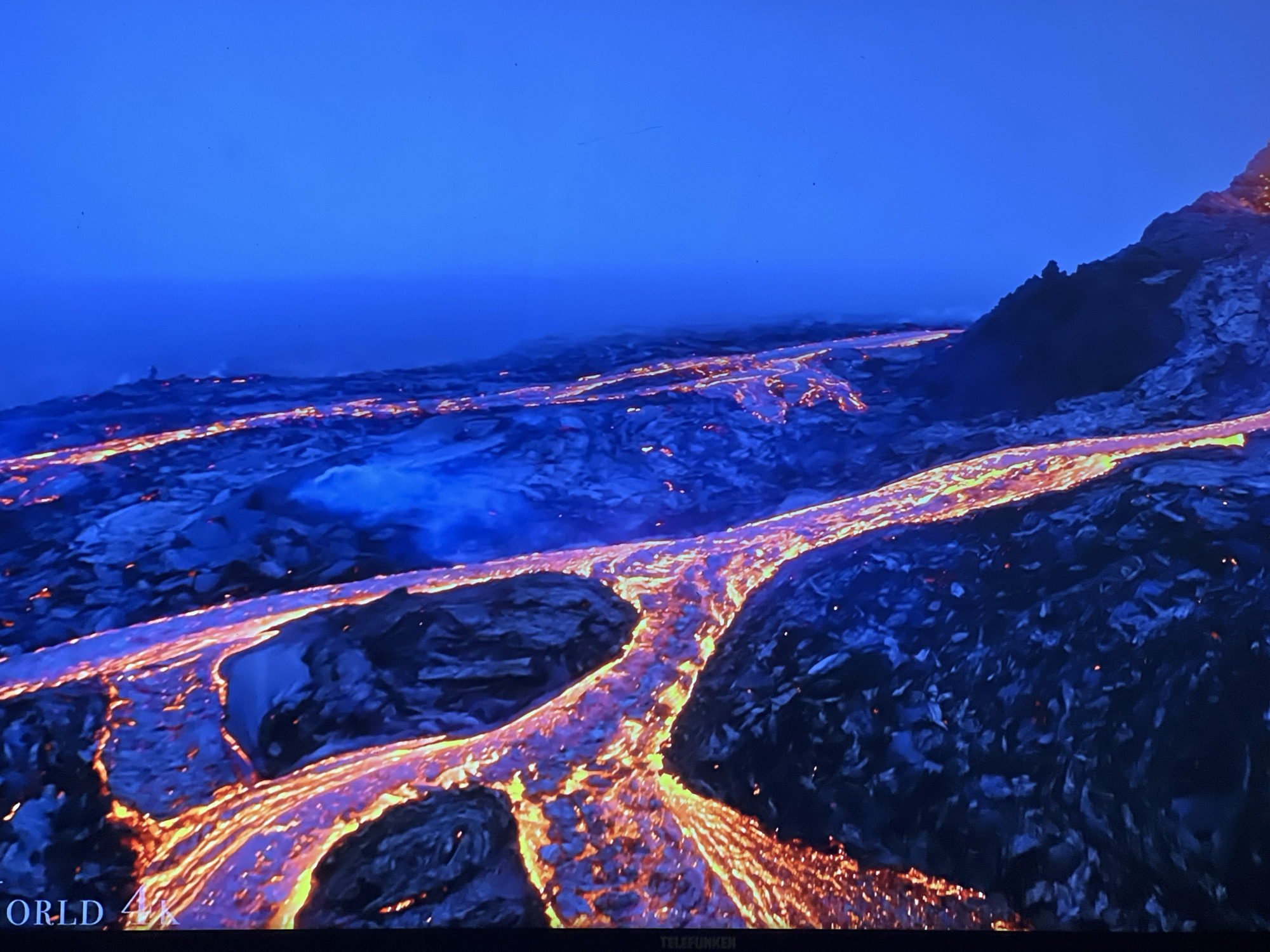 The image captures a vast, dark landscape illuminated by glowing rivers of lava. The lava flows, a vibrant orange and yellow, snake across the dark terrain, creating a stark contrast with the deep blue of the twilight sky and the surrounding volcanic rock. In the distance, a faint plume of smoke or steam rises, hinting at the ongoing volcanic activity. The foreground of the image shows more of the solidified lava, dark and textured, with small embers or glowing fragments scattered throughout. At the very bottom of the frame, text overlays are visible, reading "ORLD 4K" on the left and "TELEFUNKEN" on the right, suggesting this is a screen display of a high-definition video. The scene does not depict any elements related to the city of Sofia, Bulgaria.