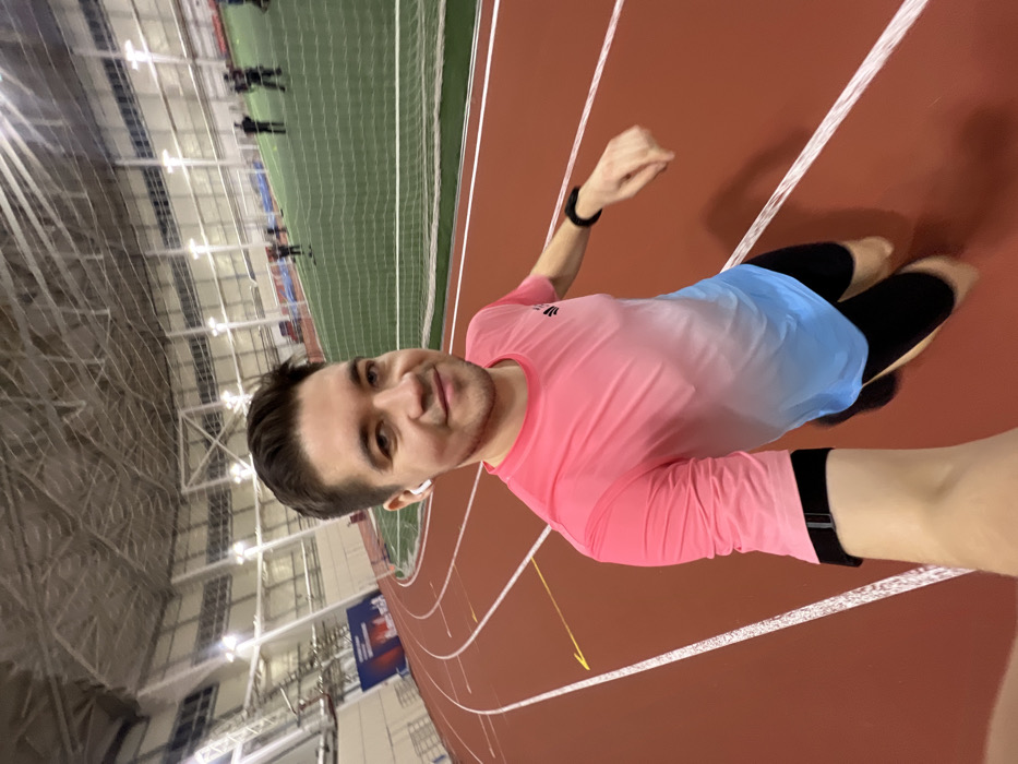 A man smiles in a selfie while jogging on an indoor track. He wears a pink and blue ombre long-sleeved shirt, black leggings, and a fitness tracker. The indoor track is red with white lines, and a green netted area borders the track. Overhead, the structure of the indoor facility is visible with bright lights. Other figures are dimly visible on the opposite side of the track.