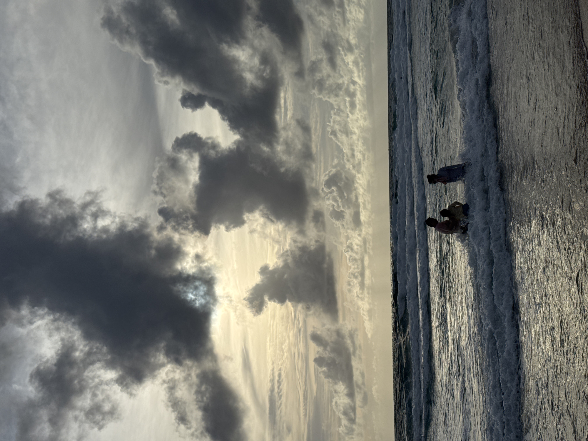 A serene view of the ocean with a family enjoying the water. The sun is setting, casting a warm glow on the clouds and the waves. The ocean is calm, with gentle waves lapping at the shore. A family of four, two adults and two children, are standing in the water, playing and enjoying the sunset. The location is Karon Beach, Karon, Thailand.