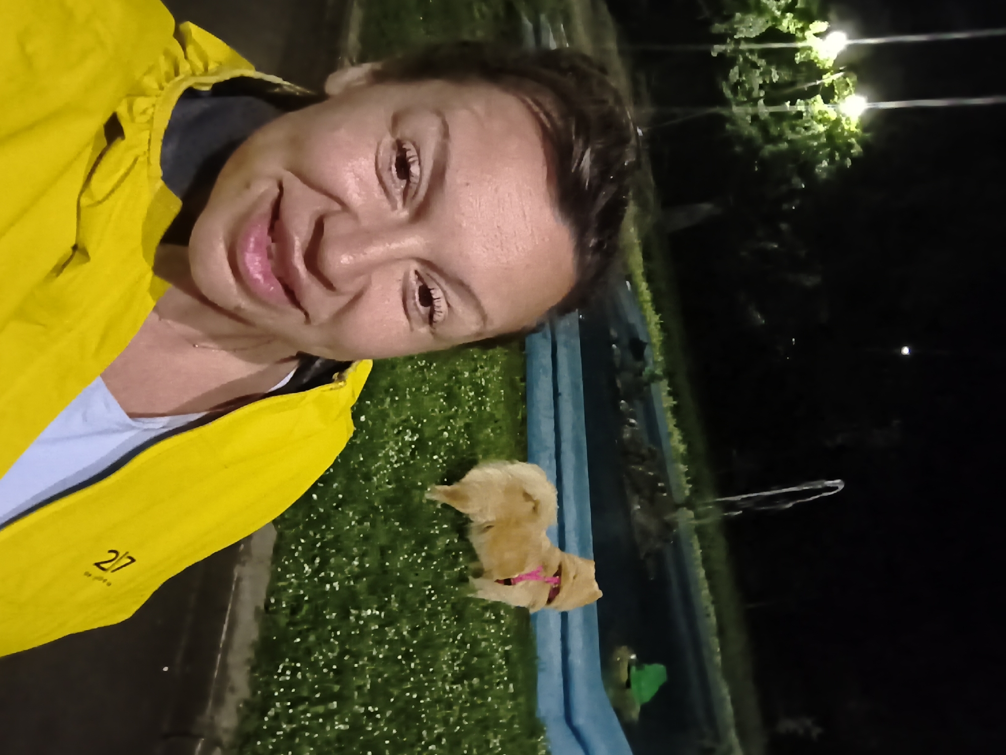 A woman wearing a yellow jacket and a white shirt is taking a selfie with a small dog wearing a pink collar at night. They are near a blue-sided body of water surrounded by greenery. Overhead, there are streetlights. "2/7" is on the jacket. The location is Varna, Bulgaria.