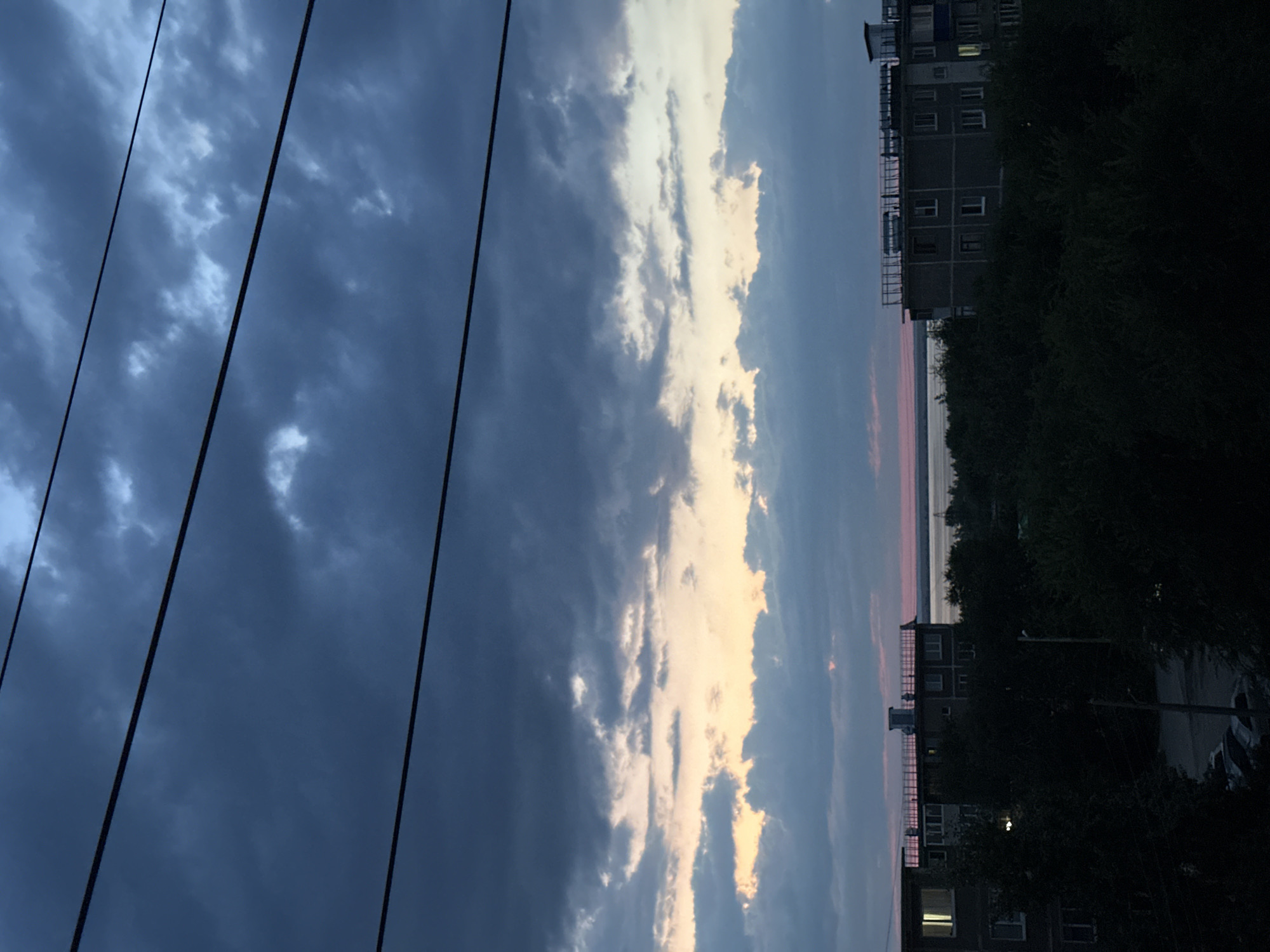 The image depicts a twilight view of Energetik, Russia, with dark, brooding clouds dominating the sky, intersected by three parallel power lines. Below the clouds, a band of soft, peachy light suggests the remnants of sunset over a distant body of water. In the foreground, silhouetted against the fading light, are the dark shapes of trees and apartment buildings. One building on the left has illuminated windows, indicating occupancy. The scene is peaceful and somewhat melancholic, characteristic of the end of the day in a residential area.