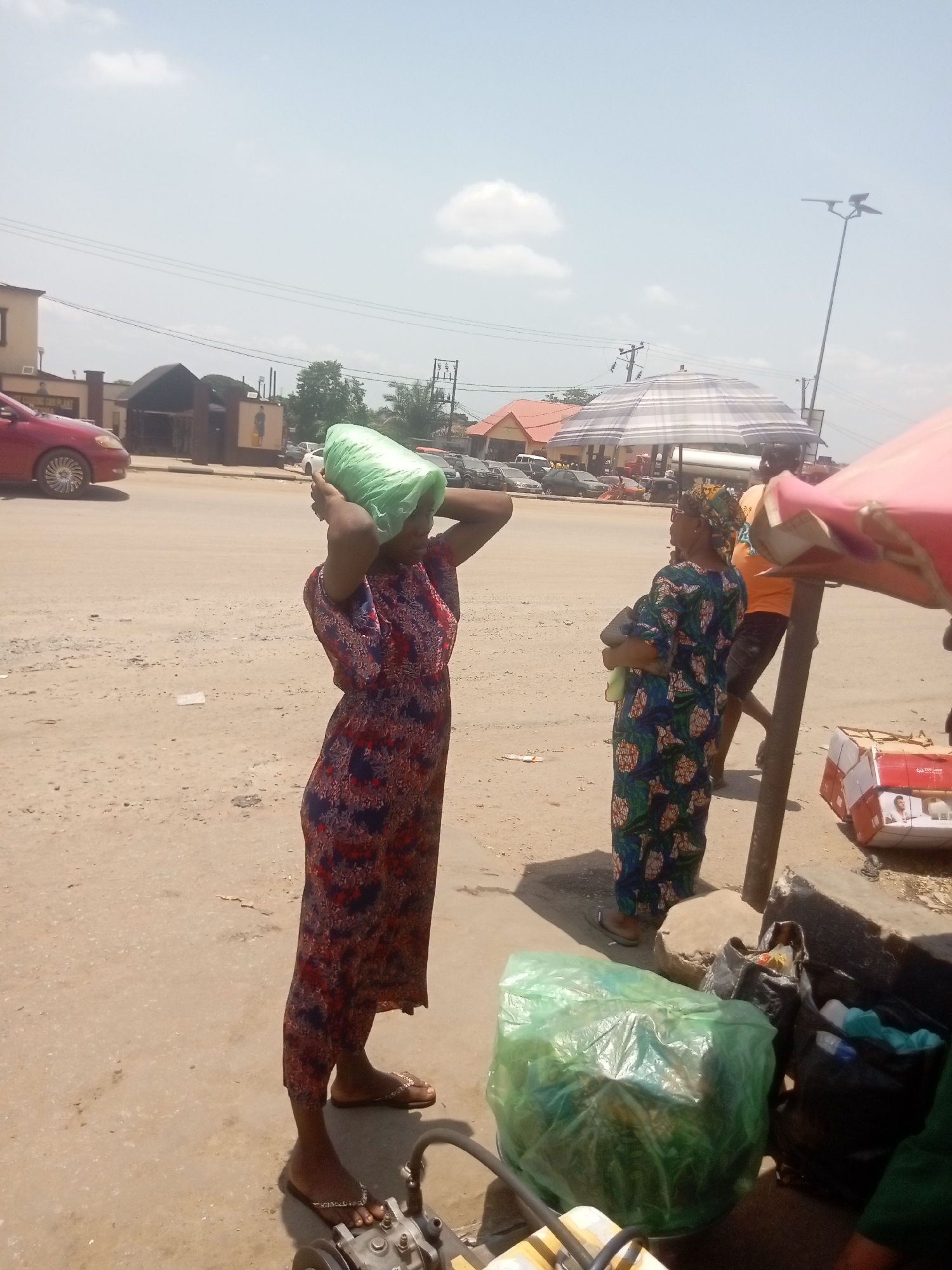 The image depicts a vibrant outdoor street scene, likely in a market or bustling area of Onitsha, Nigeria, on a bright, sunny day with a clear sky and scattered clouds. The ground is dusty and unpaved, indicative of a well-used thoroughfare.
The primary subjects are two women and a partially visible man. In the foreground, a woman stands facing slightly away from the viewer, elegantly balancing a large, bright green plastic bag on her head. She wears a long, flowing dress with an intricate pattern of red, blue, and white, paired with simple open-toed sandals. To her right, further back, another woman is seen in profile, wearing a green and blue patterned dress and a headwrap. She carries what appears to be a bundle or possibly a baby in her arms, looking towards the right side of the frame. A man in an orange shirt is partially visible behind her.
The scene is rich with contextual details. A large, grey and white striped umbrella provides shade in the middle ground, suggesting a stall or gathering point. To the right, part of a red or pink umbrella is visible, under which several cardboard boxes are stacked on the ground. One box has the word "GOLD" and a person's image, while another shows partially legible text like "SAY" and "POWER." Closer to the foreground, another large green plastic bag sits on the ground, along with several black plastic bags. In the immediate foreground, a metallic object, possibly part of a small engine or machine, rests on the ground. A red car is visible on the far left, either parked or passing by. Power lines crisscross the sky, and various buildings with local architectural styles line the background. Faint text, "HYGIENE OASIS," is visible on a building in the far left background.
The overall activity suggests daily life in a lively Nigerian street or market, with people engaged in commerce or errands under the bright daylight.