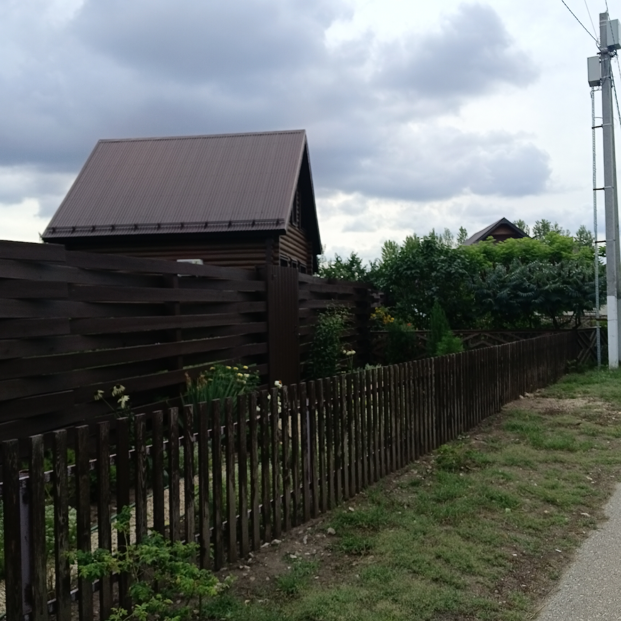 The image depicts a wooden house with a dark brown, corrugated metal roof, situated behind a dark brown wooden fence. The fence is constructed with horizontal planks of varying widths, interspersed with vertical fence posts. To the right of the house, a concrete utility pole with electrical equipment is visible, with power lines extending across the frame. The environment includes green trees and foliage, with some flowers visible behind the fence. The ground in front of the fence is covered with grass and some gravel. The sky is overcast with dark clouds. The scene is located in the village of Rozhdestveno, Russia.