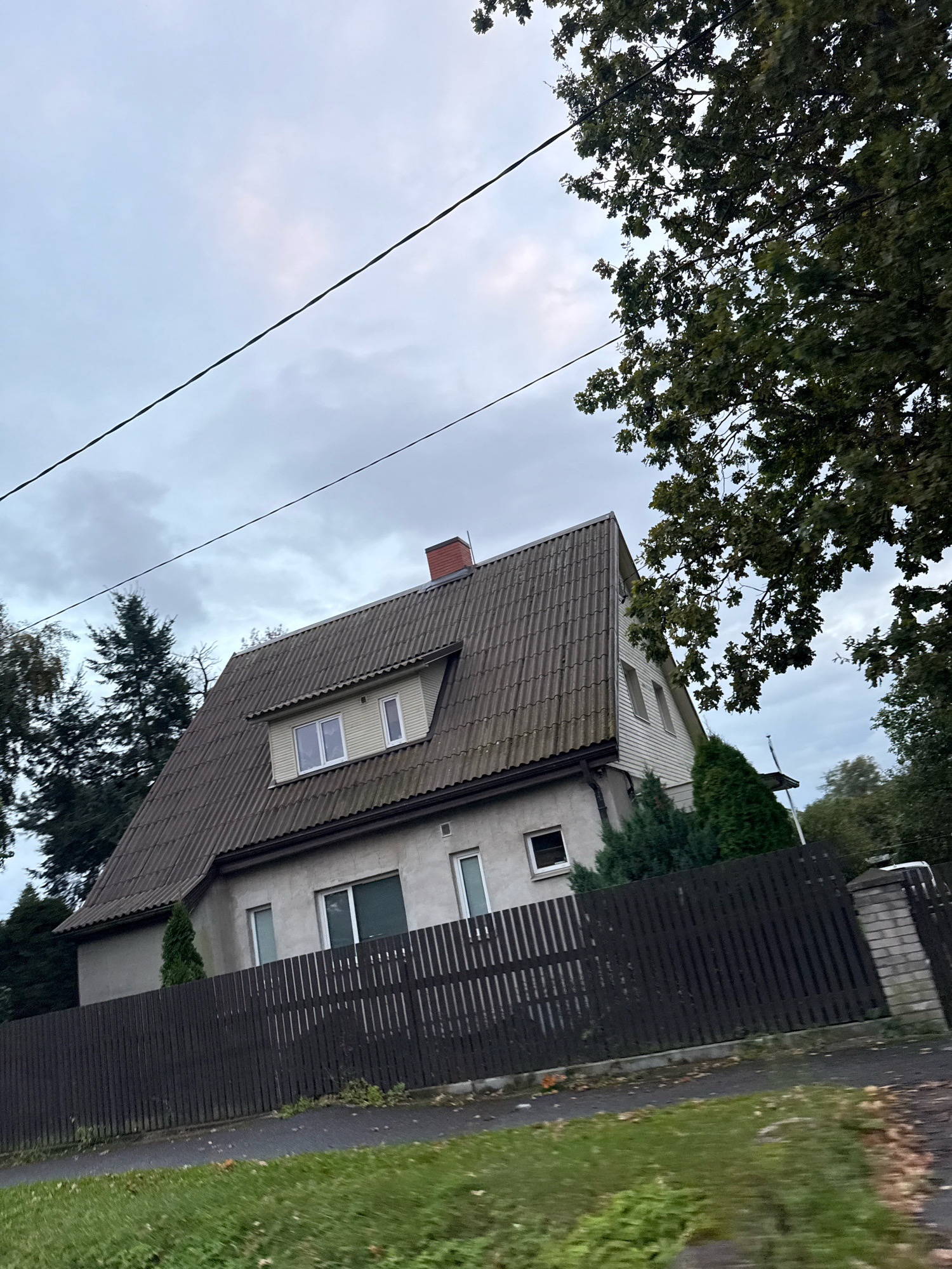 The image captures a two-story house with a steep, gray tiled roof and a dormer window on the upper floor. The house is constructed with light-colored, possibly stucco, walls and features several windows of varying sizes. A dark brown wooden fence runs across the front of the property, separating it from a grassy area and a paved path. The environment appears to be a residential area with mature trees, including evergreens and deciduous trees with green foliage, framing the scene. The sky is overcast with a cloudy, grayish hue, suggesting a temperate climate. Power lines are visible running diagonally across the upper portion of the image. The scene is static and appears to be taken from a street-level perspective, possibly from a moving vehicle due to the slight motion blur. The overall impression is of a suburban or semi-rural setting. While the image depicts a house, it does not provide specific details that would definitively place it within Tallinn, Estonia.
