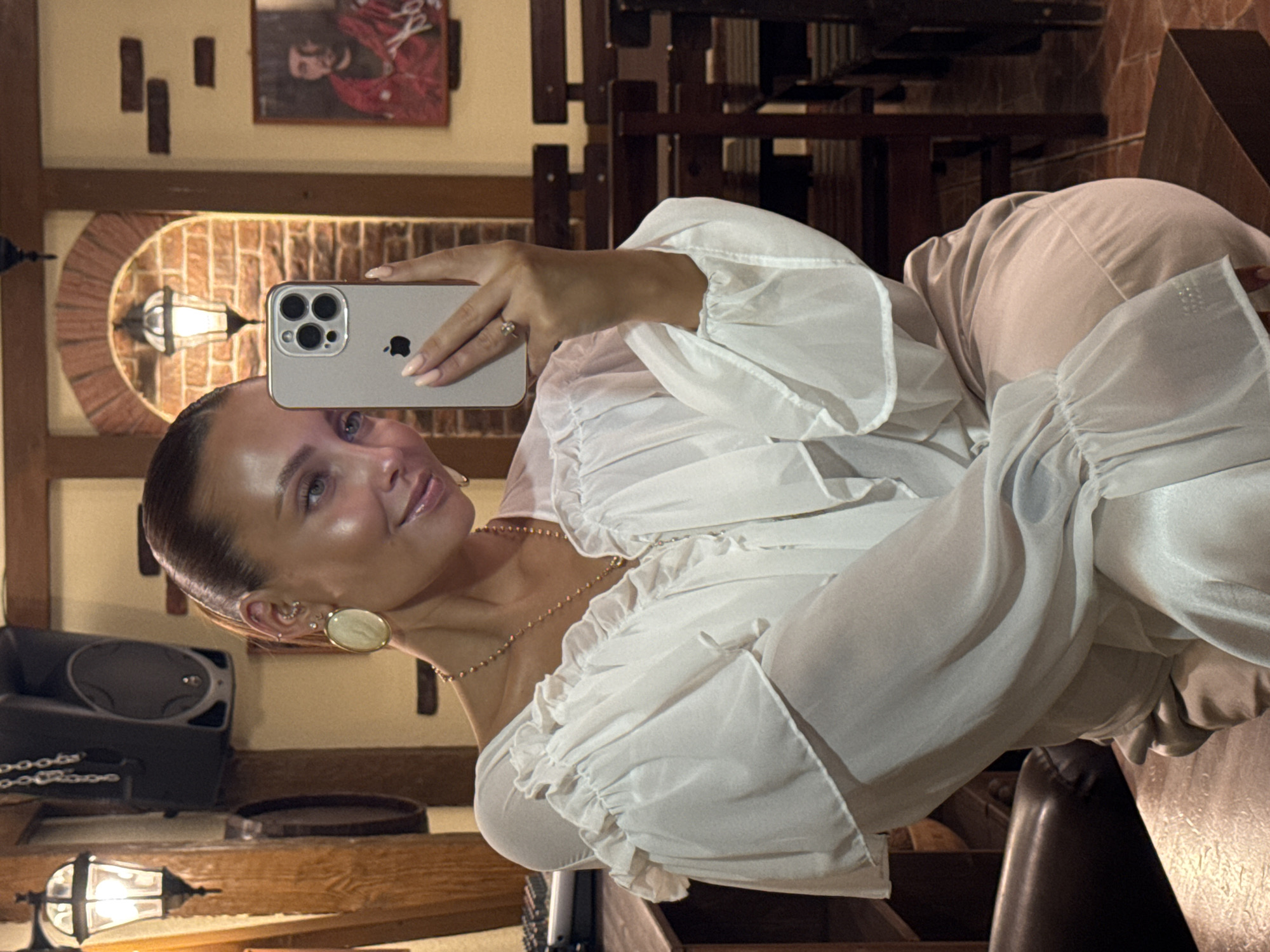 A young woman with her hair pulled back in a ponytail takes a selfie in a dimly lit establishment. She is wearing a sheer white blouse with ruffled sleeves and a pearl necklace. She is holding an iPhone in her right hand. The background shows wooden booths and tables, along with a painting on the wall. The location is identified as Chaikovsky, Russia.