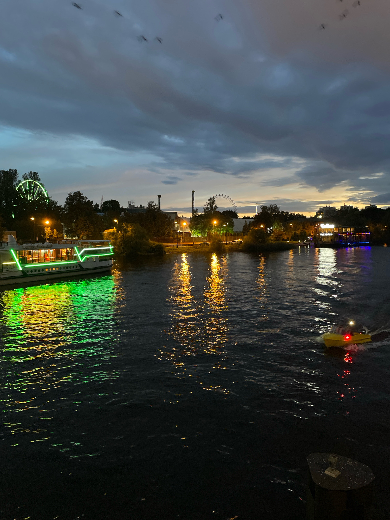 The image captures a twilight scene on the Neva River in Saint Petersburg, Russia. A large passenger boat, illuminated with vibrant green lights, is docked on the left side of the frame, its reflection shimmering on the dark water. Across the river, the city skyline is visible, featuring a Ferris wheel on the left, and various buildings with illuminated windows, some appearing to be restaurants or cafes. The sky is filled with dramatic, dark clouds, hinting at the end of the day or an approaching weather change. A small yellow motorboat, with red lights flashing, is moving across the river on the right side, creating a wake. The water is choppy, reflecting the lights from the city and the boats, creating streaks of yellow and green light. There are a few indistinct dark shapes in the sky, possibly birds or debris. The overall impression is one of a bustling urban waterfront at dusk, with the city lights beginning to dominate the darkening natural light.