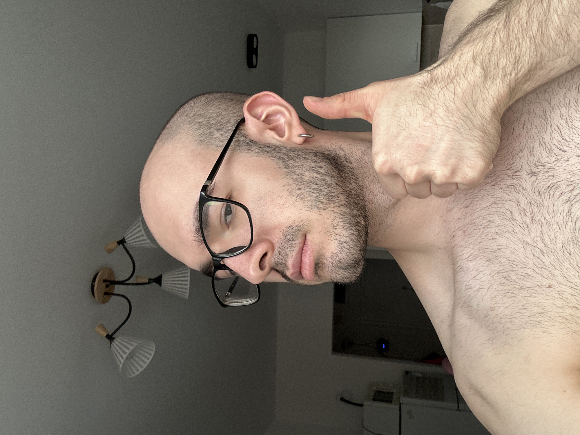 A man with a shaved head, glasses, and a beard is posing. He has a silver hoop earring in his left ear and is pointing his finger at it. He is shirtless, exposing his chest and shoulder hair. A ceiling lamp fixture is visible in the background on a gray wall. There is a cabinet and television in the background. The location in the city of Lyubertsy, Russia is unknown.