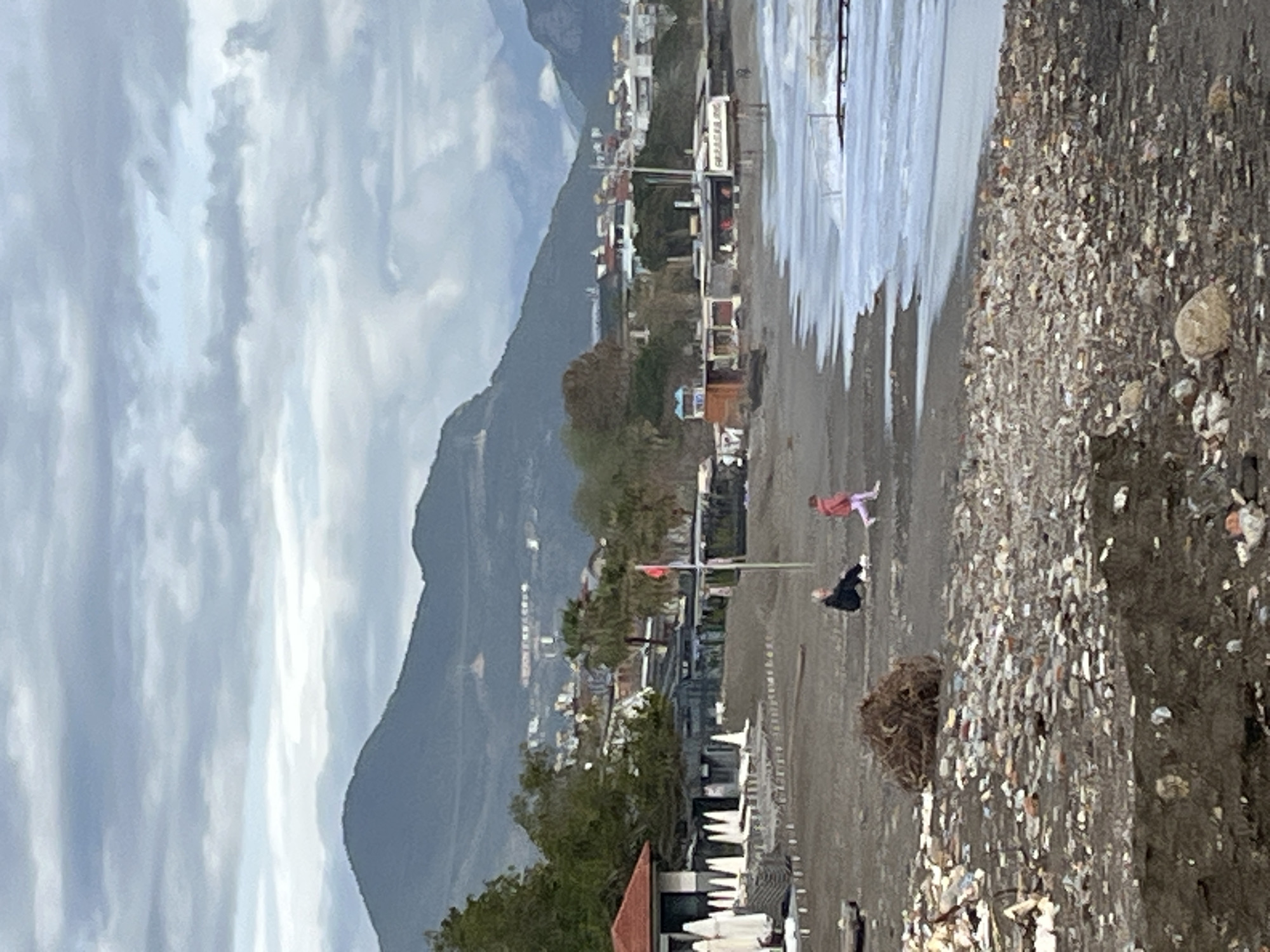 The image depicts a coastal scene in Alanya, Turkey, characterized by a beach with a mix of sand and pebbles, a distant mountain range, and a cloudy sky. In the foreground, a pile of mixed debris lies on the beach. Two figures, one dressed in pink and the other in black, are present on the beach, seemingly engaged in some activity or interaction. The background shows a cityscape nestled against the mountains, with buildings and infrastructure visible. A flag is situated on the beach. Beach chairs are stacked in the vicinity. The sea exhibits a glassy surface.