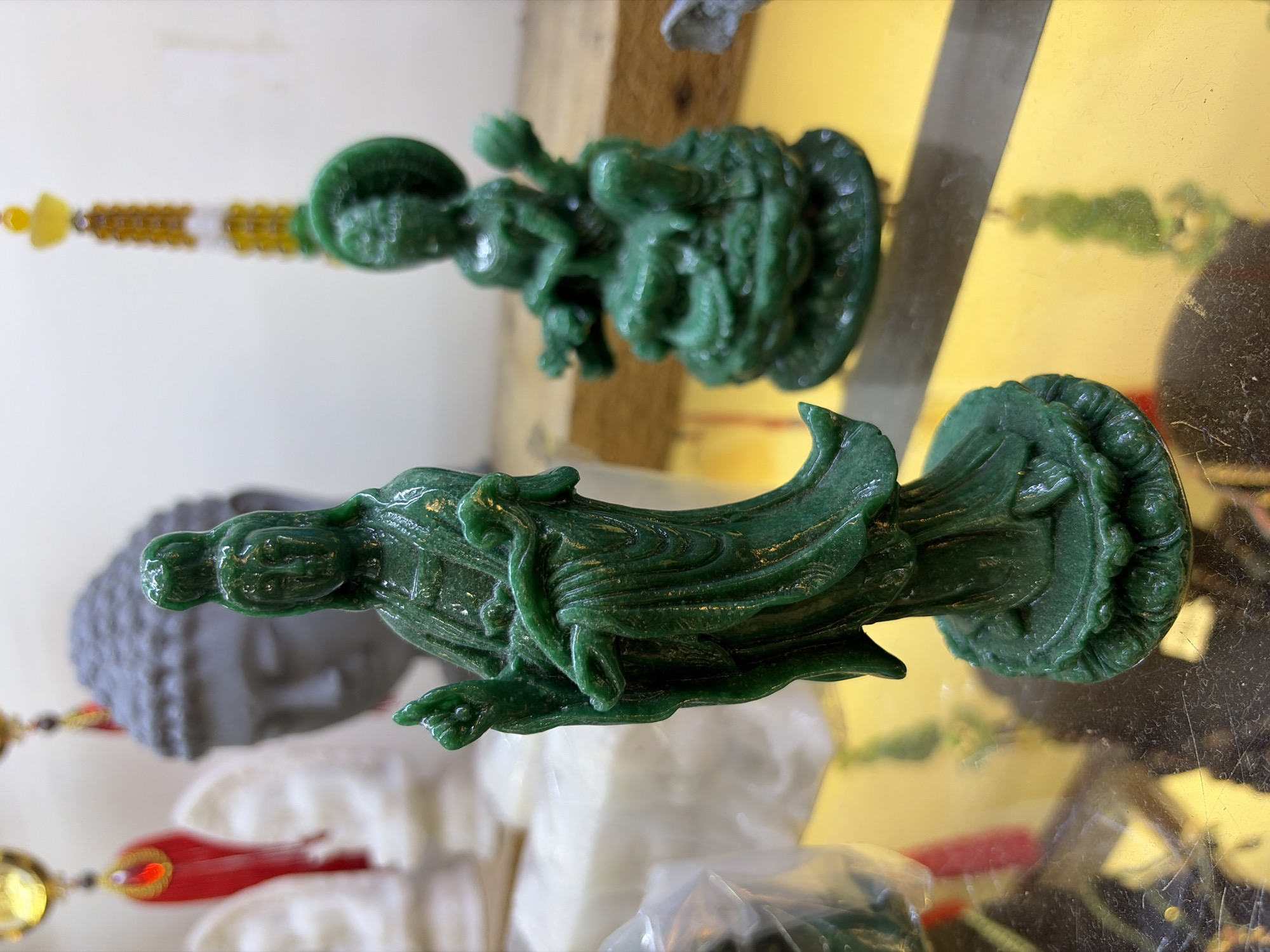 The image depicts a close-up, eye-level view of two small, green, carved figurines on a reflective, yellow surface. The figurine in the foreground is standing, with one hand raised in a gesture of blessing or teaching. It is adorned with flowing robes. Behind it and slightly to the right, a second, smaller figurine is seated on what appears to be a coiled serpent. The background is blurred, showing hints of other objects, including what looks like the head of a Buddha statue and some white, translucent forms. The overall scene suggests a display of religious or decorative items. The location is not identifiable from the image itself and is not specified in the provided context.