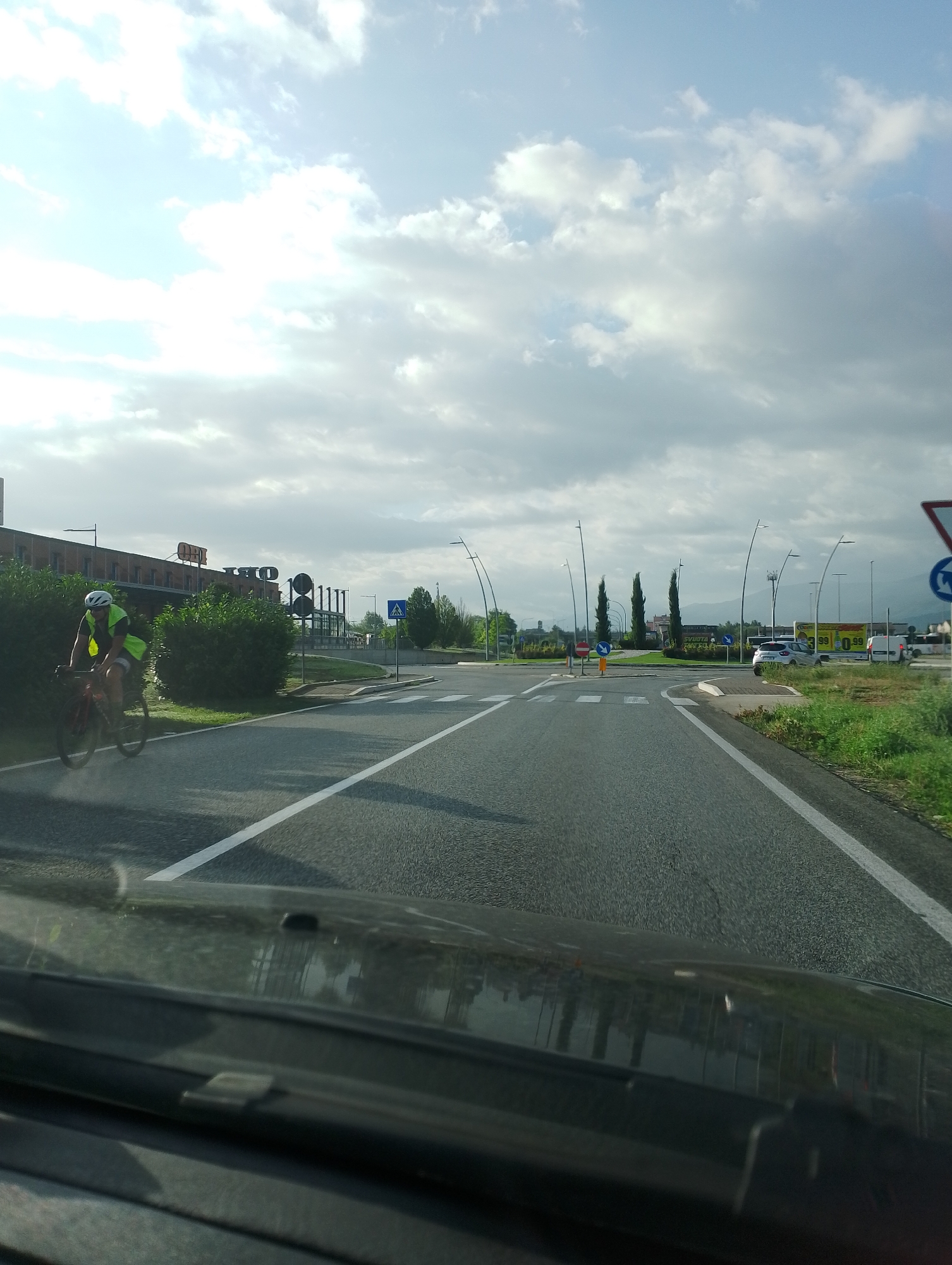 The image captures a view from a vehicle, looking forward onto a street with a cyclist on the left side. The cyclist is wearing a bright yellow vest and a white helmet, riding a red bicycle. In the background, a building with a sign that appears to read "OLIO" is visible. The street has white lane markings and a pedestrian crossing. Beyond the street, there are trees, streetlights, and a sign indicating a roundabout. The sky is partly cloudy. The scene appears to be on a road in Avezzano, Italy, likely near a commercial or industrial area.