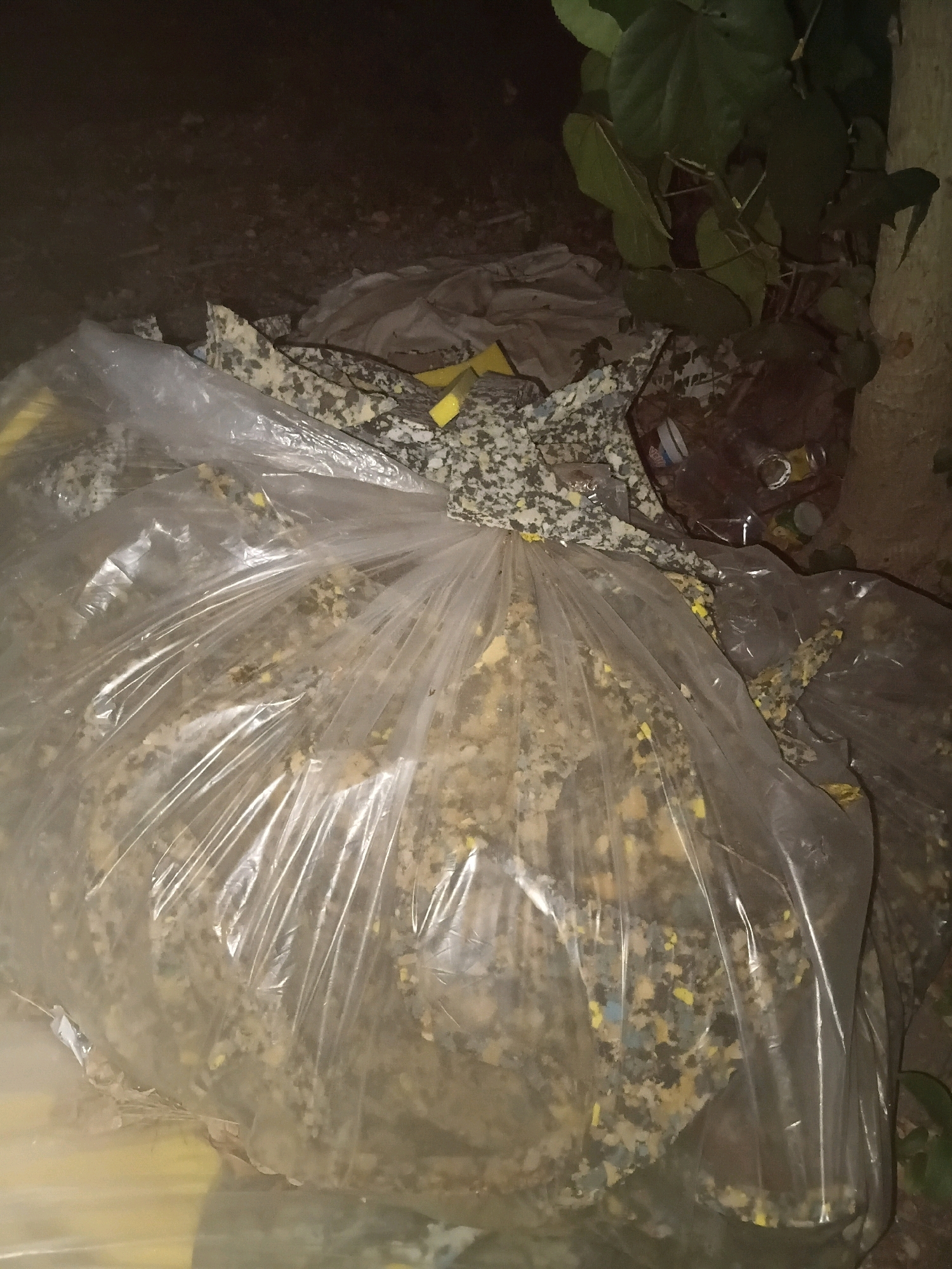 A large, clear plastic bag filled with shredded foam material is placed on the ground outdoors. The foam appears to be multi-colored, with visible flecks of yellow, gray, and white. The bag is partially open at the top, and some of the foam has spilled out. Behind the bag, there is a pile of what looks like fabric or more shredded material, and further back, there are some green leaves and dark tree bark. Several plastic bottles and cans are also visible in the background, suggesting this may be a trash or debris area. The scene is dimly lit, indicating it might be nighttime or a shaded area. The location is specified as Cikal, Indonesia.