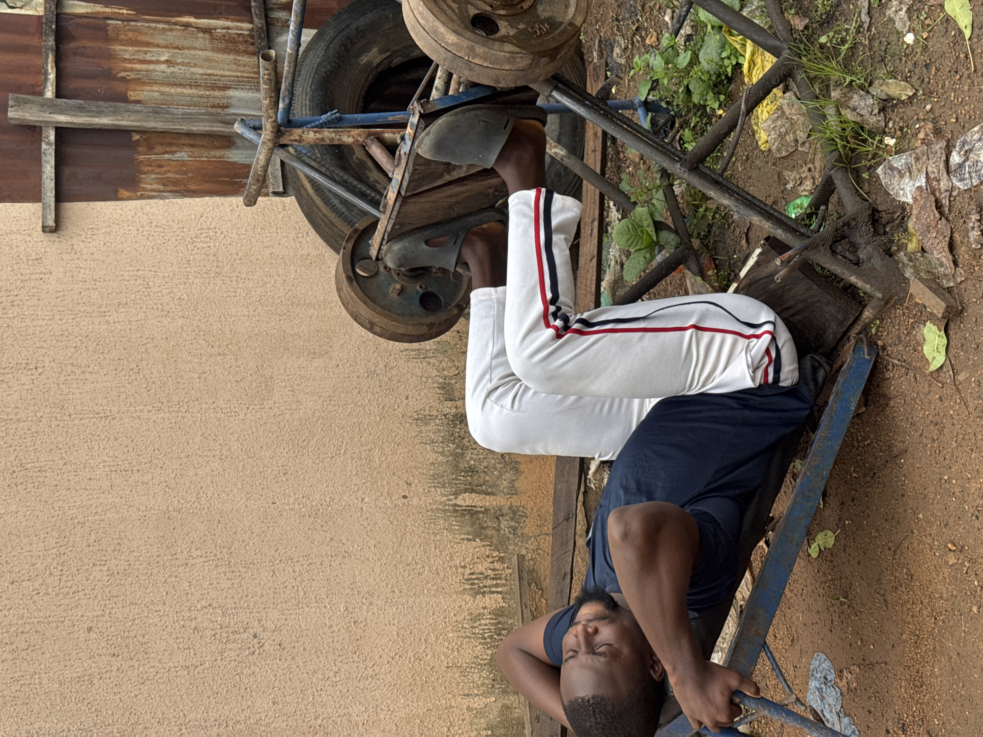 This is a vertical-format, candid, eye-level photograph taken outdoors in Akwanga, Nigeria. The scene depicts a person lying down, resting on a piece of metal equipment. The subject is wearing a dark blue t-shirt and white track pants with red and black stripes down the sides. Their eyes are closed, and their head is resting on their arms, suggesting they are asleep or deeply relaxed.
The setting appears to be an outdoor area with a rough, dirt ground scattered with debris, including leaves, plastic bags, and some vegetation. To the left, there is a textured, light brown wall. Behind the subject, a rusty metal structure with large, circular weights and a tire is visible. The lighting suggests it is daytime, possibly with overcast skies or shade. The overall impression is one of a casual, perhaps impromptu rest in an unconventional location. There is no visible text in the image.