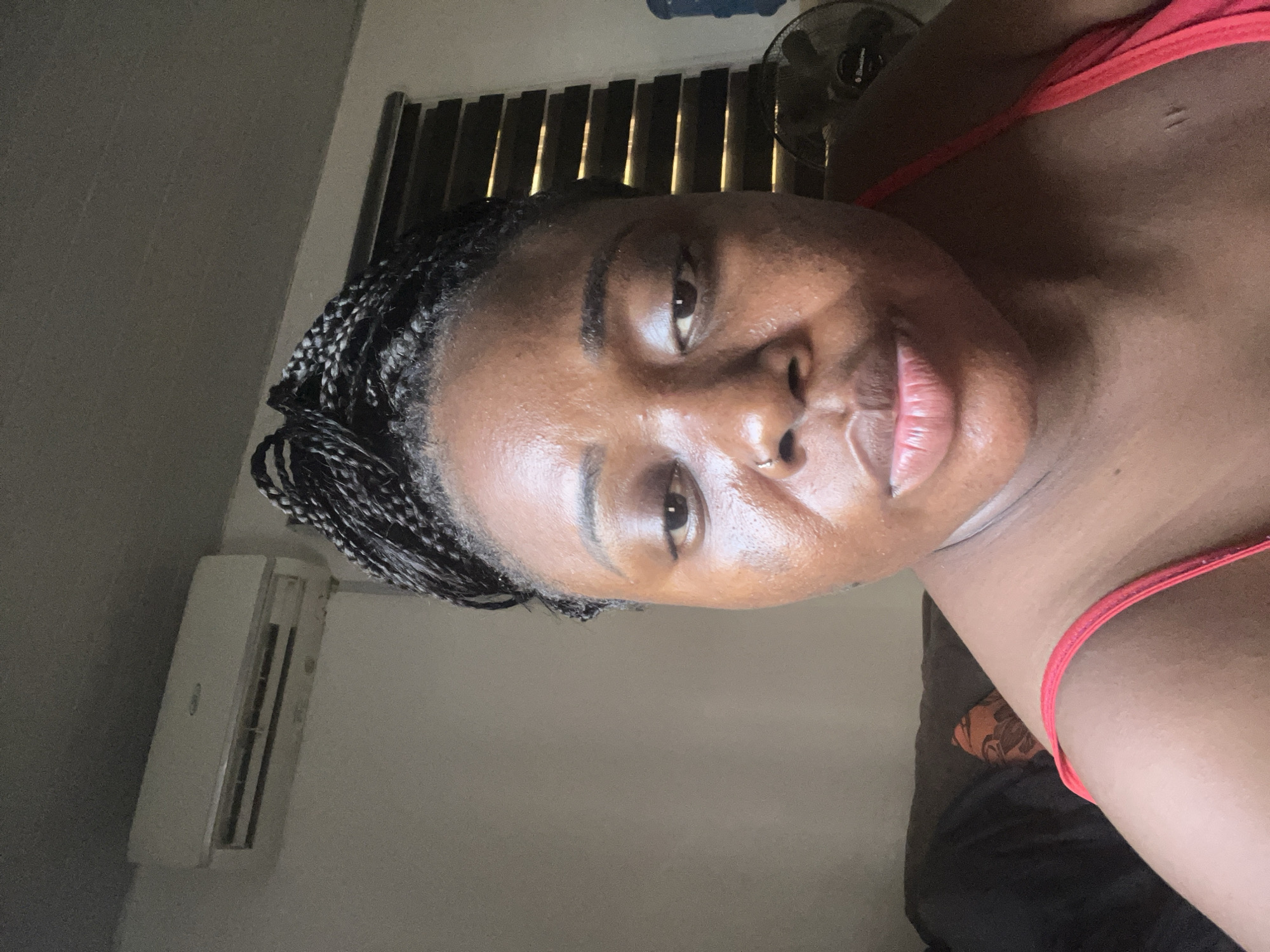 A close-up portrait of a woman with dark skin and braided hair. She is wearing a red top and appears to be indoors. The background shows an air conditioning unit, a window with blinds, and a fan. There is no information available to determine the precise location in Abuja, Nigeria.