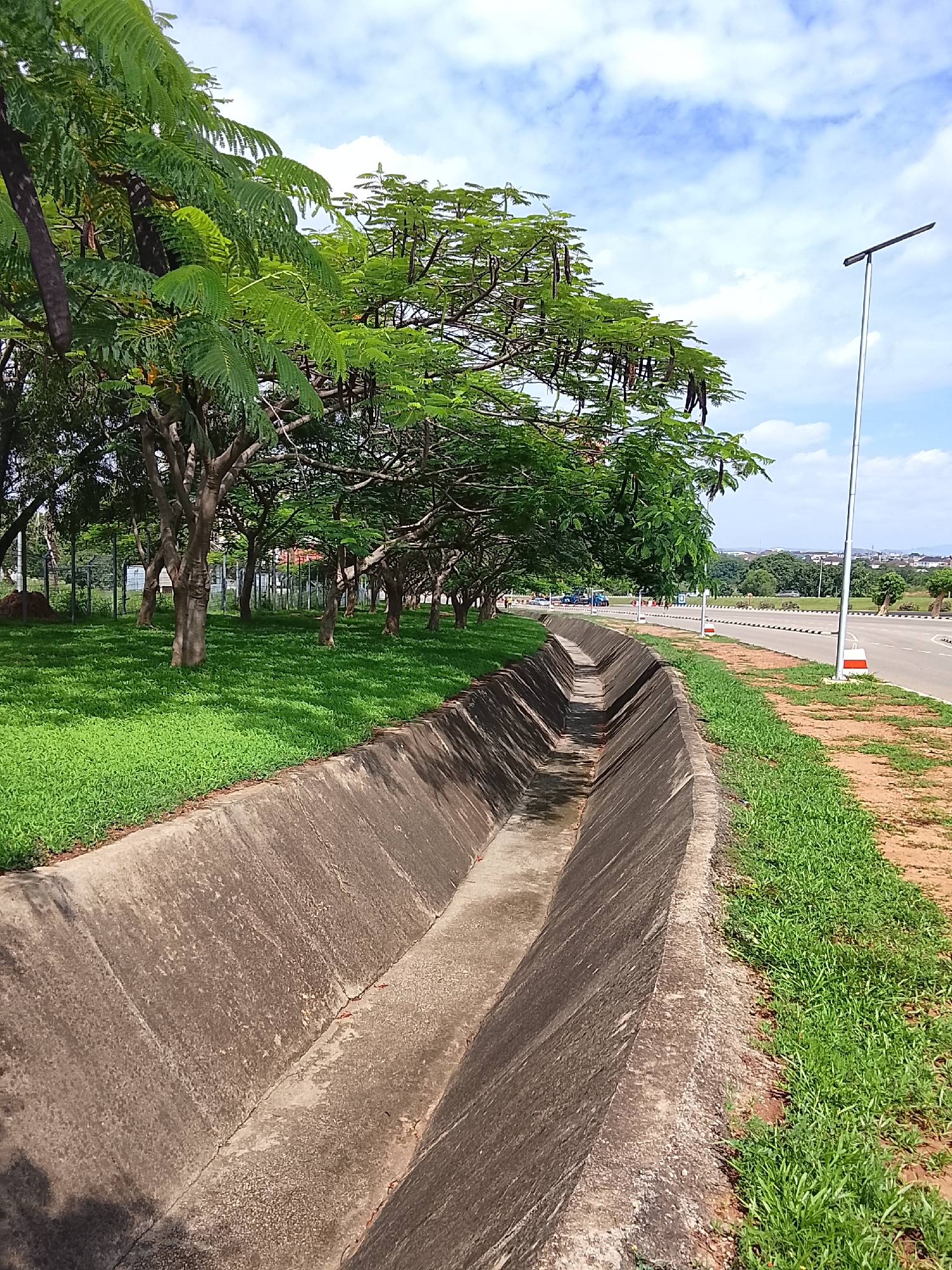 The image depicts an urban landscape in Karmajiji, Nigeria, under a bright blue sky with scattered white clouds. A concrete, V-shaped drainage ditch runs diagonally from the lower-left foreground to the mid-ground, exhibiting weathering on its surfaces. To the immediate left of the ditch, a strip of lush, manicured green grass extends beneath a line of mature trees, many of which display distinct dark, elongated seed pods hanging from their branches. A metal mesh fence is visible behind the trees. To the right of the ditch, a strip of reddish-brown dirt and sparse green grass borders a wide, multi-lane paved road. The road is marked with white lines and features white and black striped barriers along its median. A tall, slender metal pole topped with a horizontal, rectangular light fixture stands upright on the right side of the ditch in the mid-ground. In the mid-ground, several indistinct human figures and at least one blue vehicle are visible on the far side of the road. The distant background reveals outlines of buildings and rolling terrain on the horizon. The scene is brightly illuminated by direct sunlight, casting discernible shadows.