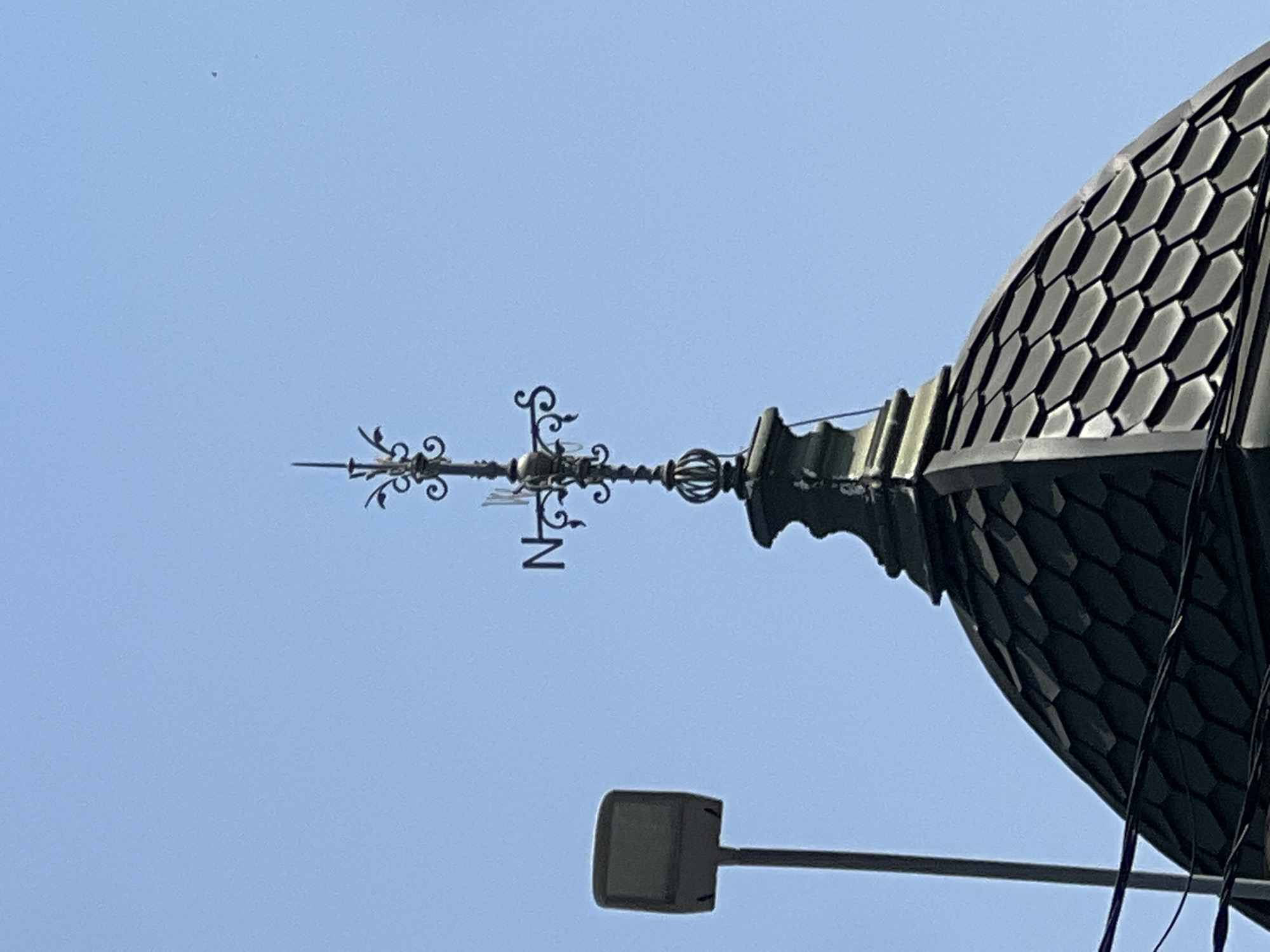 The image shows the top of a building with a dark, hexagonal-tiled dome, topped by an ornate weather vane against a clear blue sky. A street lamp with a rectangular fixture stands to the left of the dome. The weather vane has decorative metalwork and points labeled "N" for North and "S" for South. The scene is static and appears to be an outdoor shot taken from a low angle, looking up. No people or animals are visible. The image does not contain any information that would allow for the identification of its specific location in the city of Krasnodar, Russia.