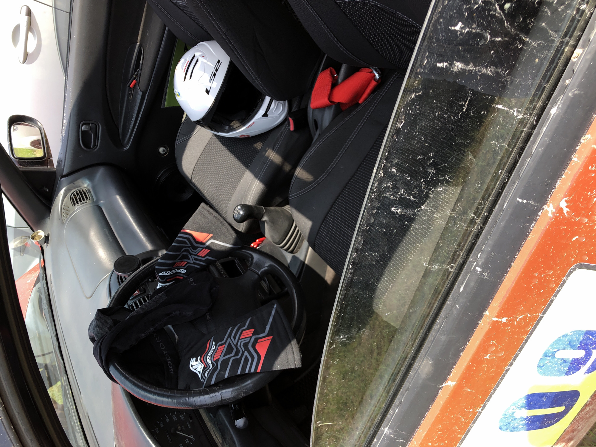 The interior of a worn two-door vehicle, likely a compact car, is depicted from an elevated side perspective, set in an unspecified outdoor location. The car's dashboard and door panels are dark grey/black and show signs of wear and dust accumulation. A black steering wheel, wrapped in a textured material, occupies the left side of the frame. Draped over the steering wheel is a black athletic top or undershirt, featuring a white and red stylized logo, the word "BODYDRY" on the left spoke, and "SPORT" with a geometric pattern on the right spoke. A manual gear stick with a black knob is positioned between the driver and passenger seats.
The passenger seat, upholstered in dark grey fabric with visible white stitching, holds a white full-face motorcycle or racing helmet displaying the "LS2" brand name and red/black stripe accents. Adjacent to the helmet, on the passenger seat and center console, a bright red folded racing harness with metal buckles is visible. The passenger door panel is dark grey/black, showing scuffs and dirt, and contains a storage pocket with what appear to be red-capped pens. A gold-colored cylindrical object is mounted on the dashboard near the windshield on the passenger side, and a circular black mount (possibly for a phone) is on the center console.
Through the dirty side window in the foreground, an orange exterior body panel of the vehicle is visible, featuring a large blue number "09" as part of a decal, also covered in dirt and specks. Beyond the vehicle's interior, through the windows, an outdoor environment is seen. The reflection in the passenger side mirror shows green foliage. Through the open passenger window, a portion of a light-colored (likely white or silver) vehicle's side, including its door handle and mirror, is visible against a bright background. Green foliage is also discernible outside the vehicle.
