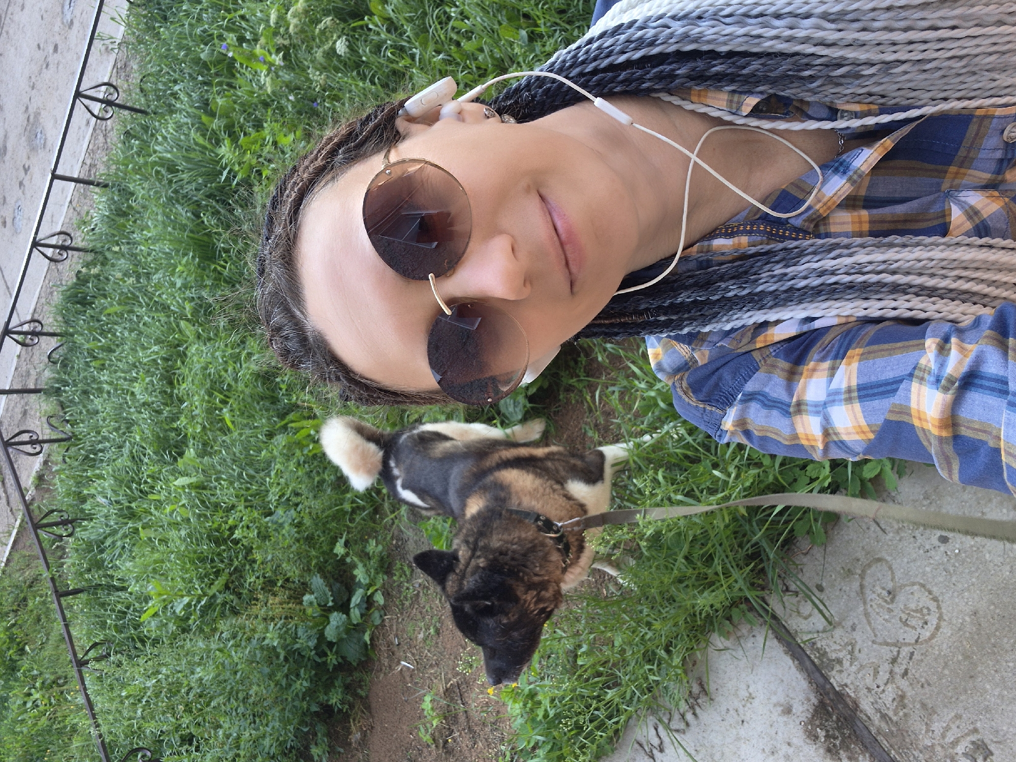 The image is a selfie taken outdoors, likely in a park or green space in Chaikovsky, Russia. In the foreground, a woman with braided hair and sunglasses is looking at the camera. She is wearing a plaid shirt and white earbuds. To her left, a dog with black, brown, and white fur is on a leash, sniffing the ground. The background consists of lush green grass and a metal railing. The ground is a mix of dirt and concrete. The lighting suggests it is daytime, possibly late afternoon, with soft shadows.