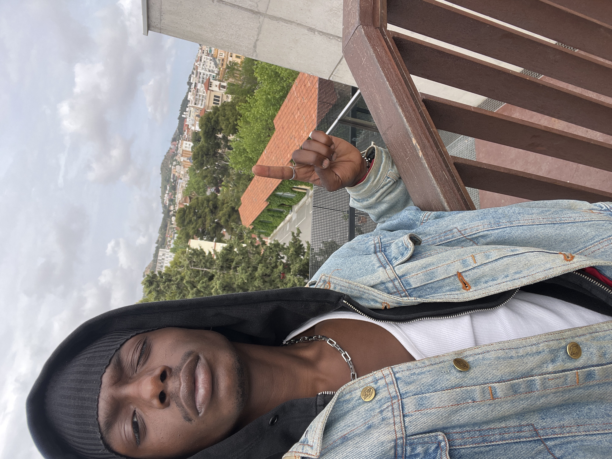 A man wearing a black beanie, black hoodie, white tank top, and light blue denim jacket is facing the camera with his eyes mostly closed and a slight smile on his face. He is extending his index finger. He is positioned on a balcony with a wooden railing. In the background is a view of Barcelona, Spain, with buildings, trees, and a cloudy sky.