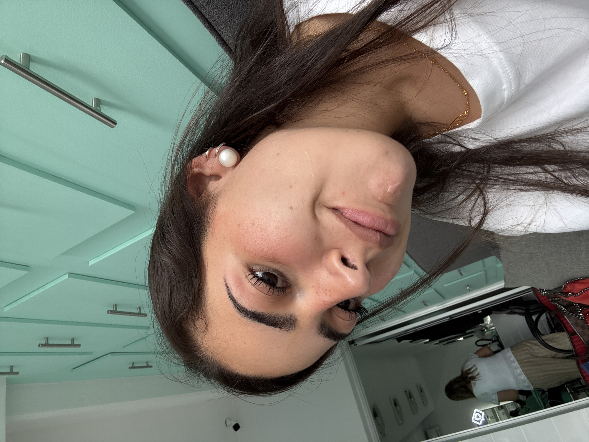 A woman's face is close to the camera, slightly turned to the side. She has dark hair, brown eyes, and fair skin. She's wearing a pearl earring and a gold necklace. Behind her, a pale green wall with silver handles suggests a bathroom or vanity area. In the reflection of a mirror, a person is visible wearing a white shirt and tan pants. There's also a red and black bag. The location is potentially San Bernardino Tlaxcalancingo, Mexico.