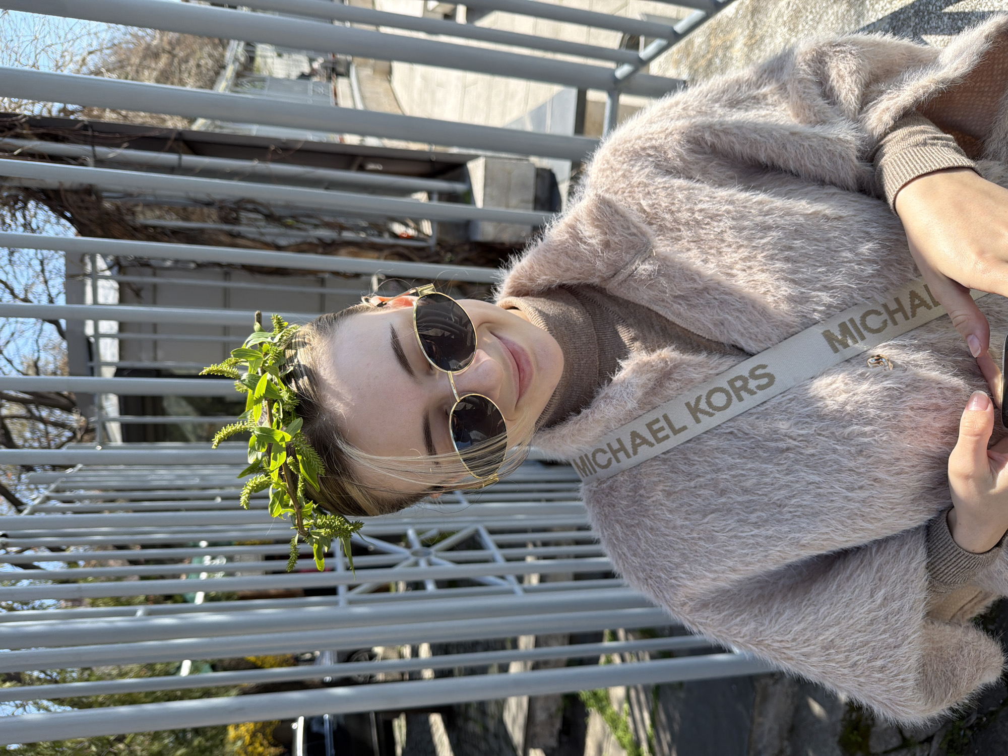 A young woman stands on a bridge in Plovdiv, Bulgaria. She wears a beige fuzzy jacket with a Michael Kors branded strap across her chest, a brown turtleneck sweater, and round sunglasses. A wreath of green leaves adorns her head. The bridge has grey metal railings. Visible beyond the bridge are trees without leaves and buildings. The day appears sunny.