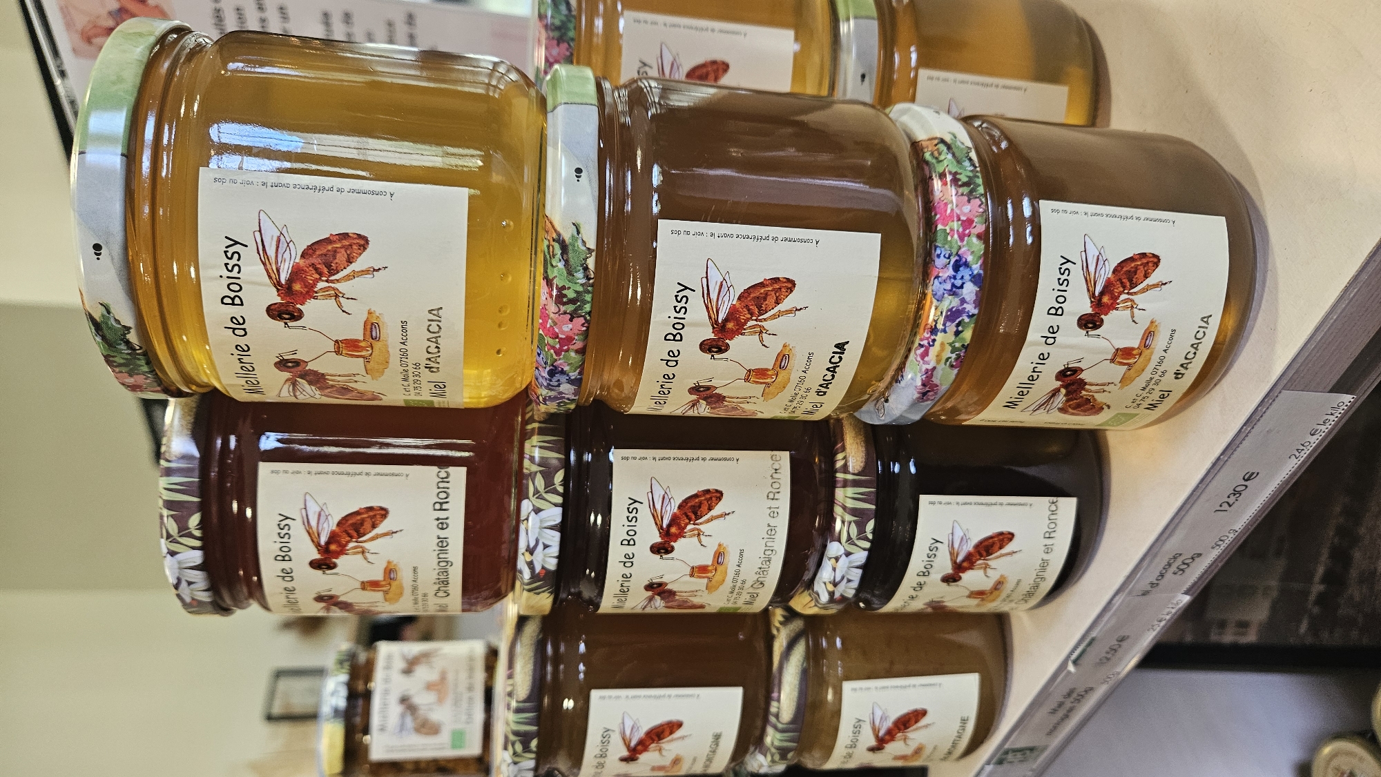 The image displays a close-up of several jars of honey stacked on a shelf. The jars are glass and have decorative lids with floral patterns. Each jar features a label with an illustration of two bees and text identifying the brand as "Mellerie de Boissy" and the type of honey, such as "Miel d'Acacia" or "Châtaignier et Ronce". Some labels also include an address and phone number, "C. r.c. Molle 07160 Accons" and "04 75 29 30 66". In the foreground, a shelf with price tags is visible. One price tag reads "12,30 €". The background is blurred but suggests an indoor retail environment. There are no people or animals visible in the image. The scene appears to be a display of honey products in a shop, likely in or near the location mentioned on the labels, Accons, which is in the Ardèche department of France. Saint-Sauveur-de-Montagut is a commune in the Ardèche department, located approximately 30 kilometers north of Accons, suggesting this shop could be a local producer or retailer in that region.