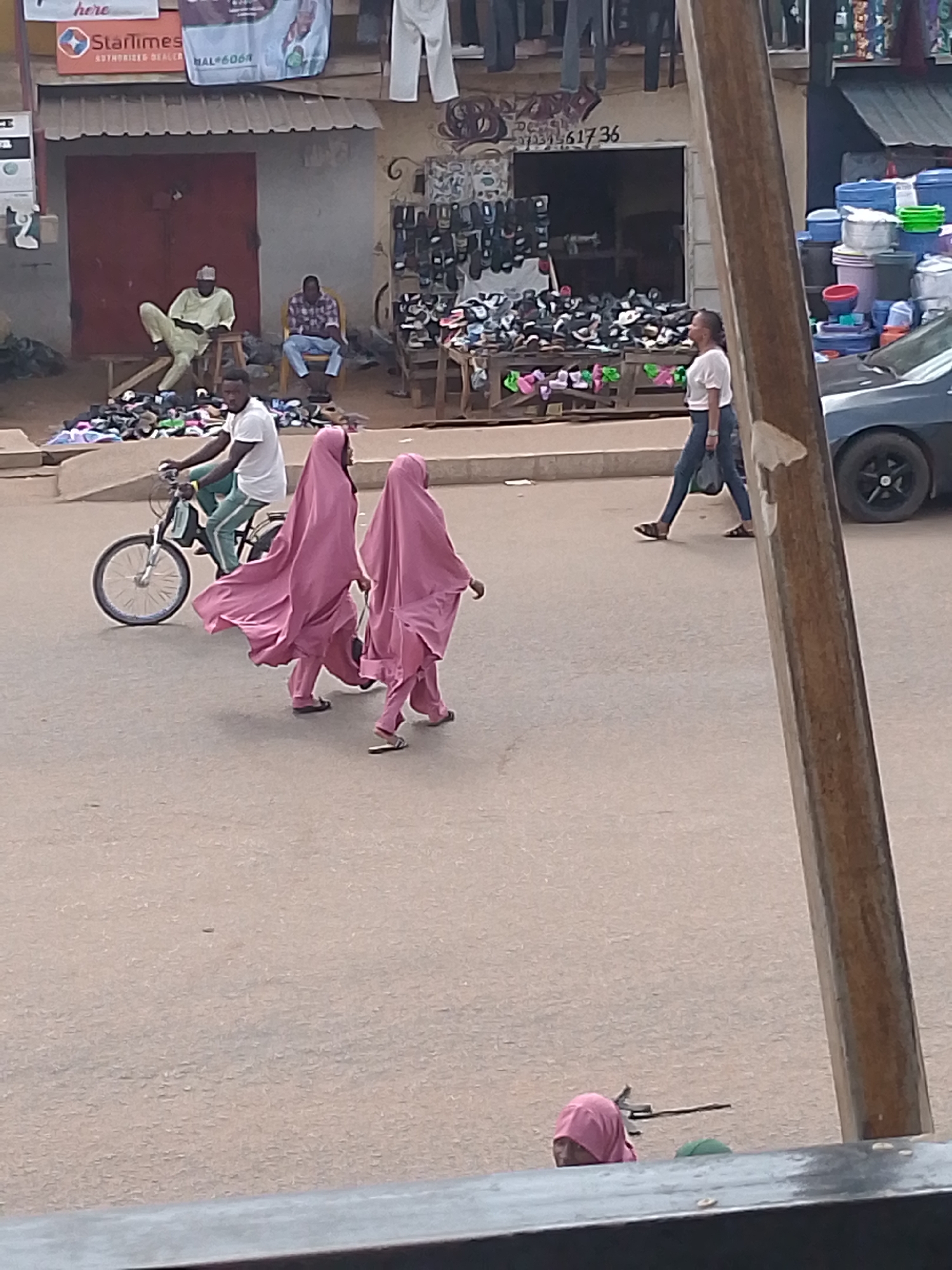 The scene is set in a city market area in Bukuru, Nigeria. Two women are walking along a street. They are wearing light pink hijab coverings. A man on a bicycle is riding behind them. There is a woman walking across the road from the opposite direction. A dark gray sedan car is parked on the side of the street next to her. Several people are seated on chairs in front of buildings. Two men are sitting in front of a building with a red metal door, surrounded by shoes. A storefront displays an array of shoes for sale. Stacks of plastic containers are piled up at a building on the right side of the scene. A pole is visible in the foreground.