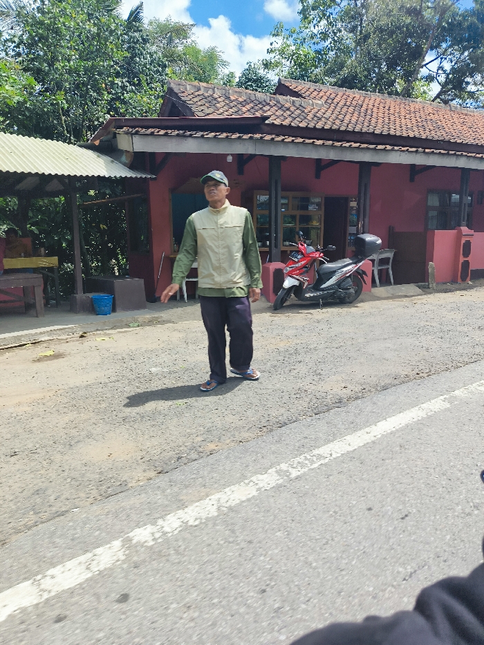 The image shows a man standing in front of a red-painted building with a tiled roof, in Cipatat, Indonesia (latitude: -6.785, longitude: 107.435). He is wearing a tan vest and green pants, a blue cap, and is looking to the left. He is standing in front of a small, red motorcycle. The man is standing on a paved road with a white line in the middle, running parallel to the building. There is a grassy area in the front, to the left of the road, that is overgrown with vegetation. In the background, to the left of the building, there is another small structure with a tiled roof and a blue plastic container on the ground. There are trees behind the structures. The sky is mostly blue, with a few white clouds. The picture was taken from the perspective of someone driving on the road.