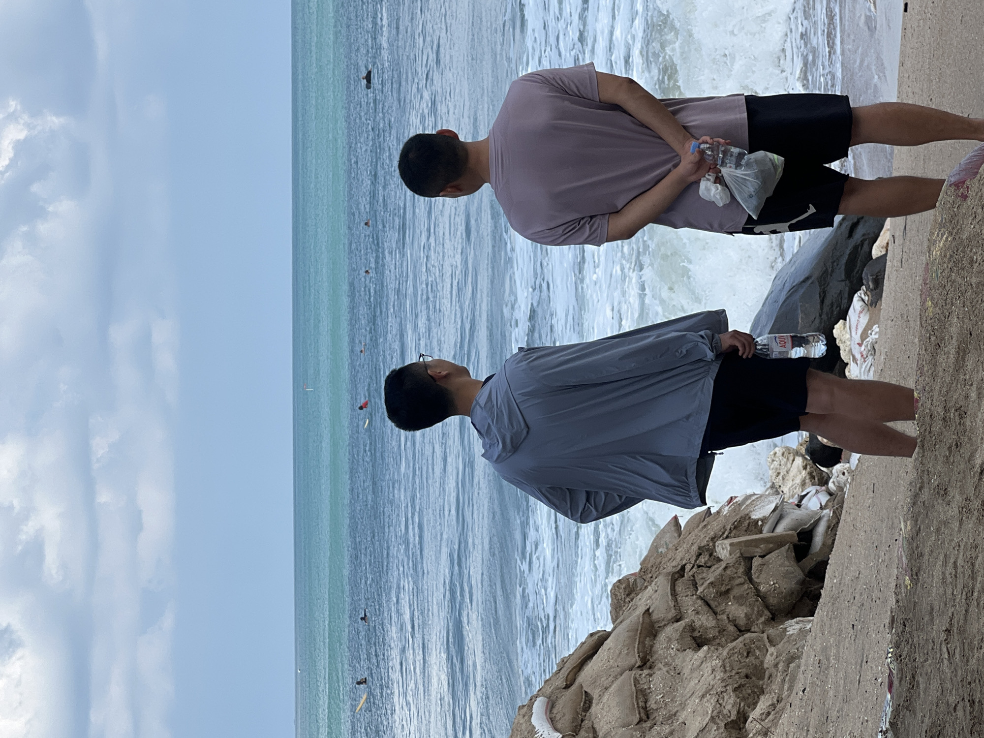 The image depicts two men standing on a sandy beach in Kuta, Indonesia, facing a choppy ocean. The man on the left wears a grey hooded jacket and black shorts, while the man on the right wears a mauve t-shirt and black shorts. Both men are holding water bottles, and the man on the right also holds a plastic bag. The background shows the ocean with several surfers in the distance. The beach is littered with debris and sandbags, suggesting erosion control measures. The sky is overcast, with light clouds. The water is a turquoise hue, transitioning to a darker blue further out. The waves are breaking onto the shore, creating white foam.