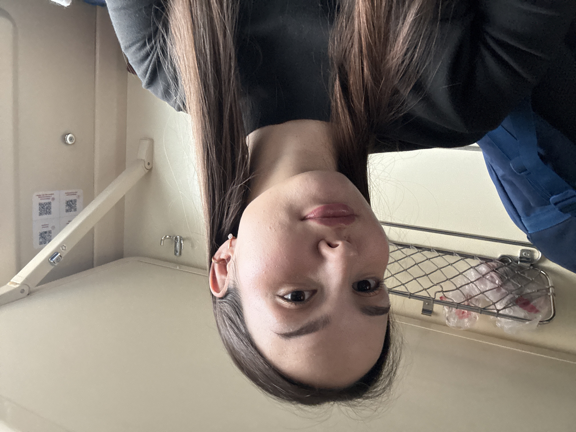 The image depicts a female individual within a train compartment, situated in Usolye-Sibirskoye, Russia. The subject, with long, straight, dark brown hair and a fair complexion, occupies the foreground, looking directly at the camera. She is wearing a black long-sleeved garment. Her right ear features a small hoop earring with a clear stone on the lobe and a gold-colored hoop earring on the helix. The background reveals beige-paneled walls typical of a passenger train. Above the subject's left shoulder, a metal mesh rack holds two clear plastic bags containing indistinct items. To her right, a set of metal coat hooks is affixed to the wall. Further right, an articulated wall-mounted mechanism, possibly for a fold-down table, is visible, along with two unreadable QR code decals. A blue backpack is partially visible on the far left side of the frame.
