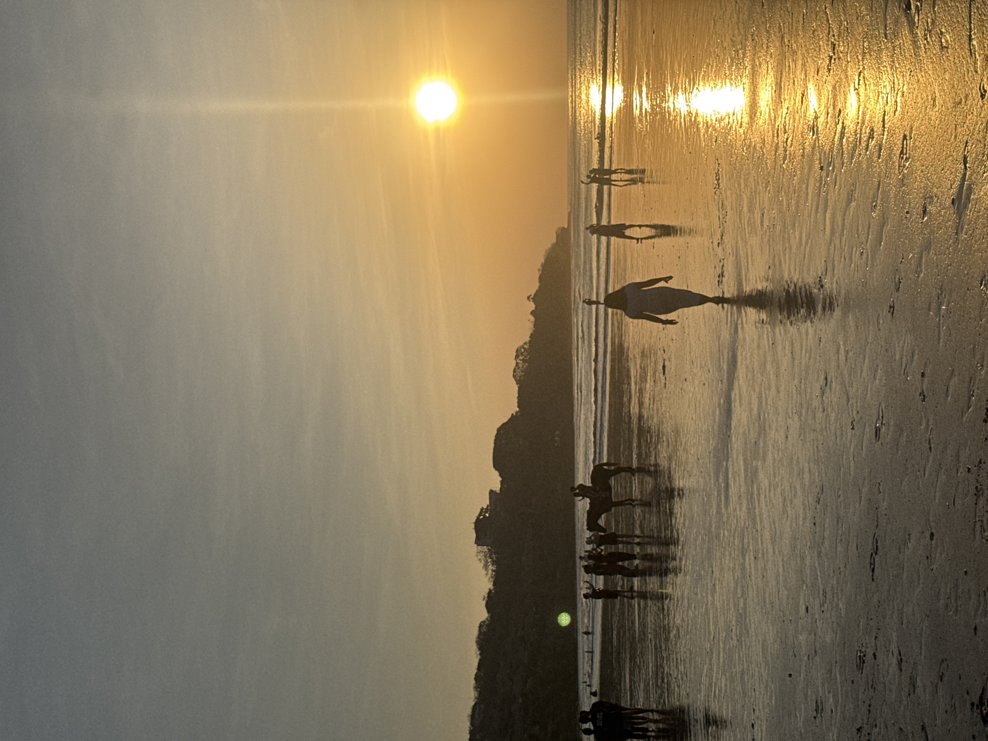 The image depicts a sunset scene at the beach in Kuta Selatan, Indonesia. Several people are visible on the wet sand, some walking and others gathered in groups. A horse with riders is also present near the water's edge. The sun, a bright orb, is setting over the ocean, casting a warm, golden glow on the sky and reflecting on the water and wet sand. The horizon is marked by a tree-covered hillside with a building structure on top. The overall atmosphere is serene and recreational.