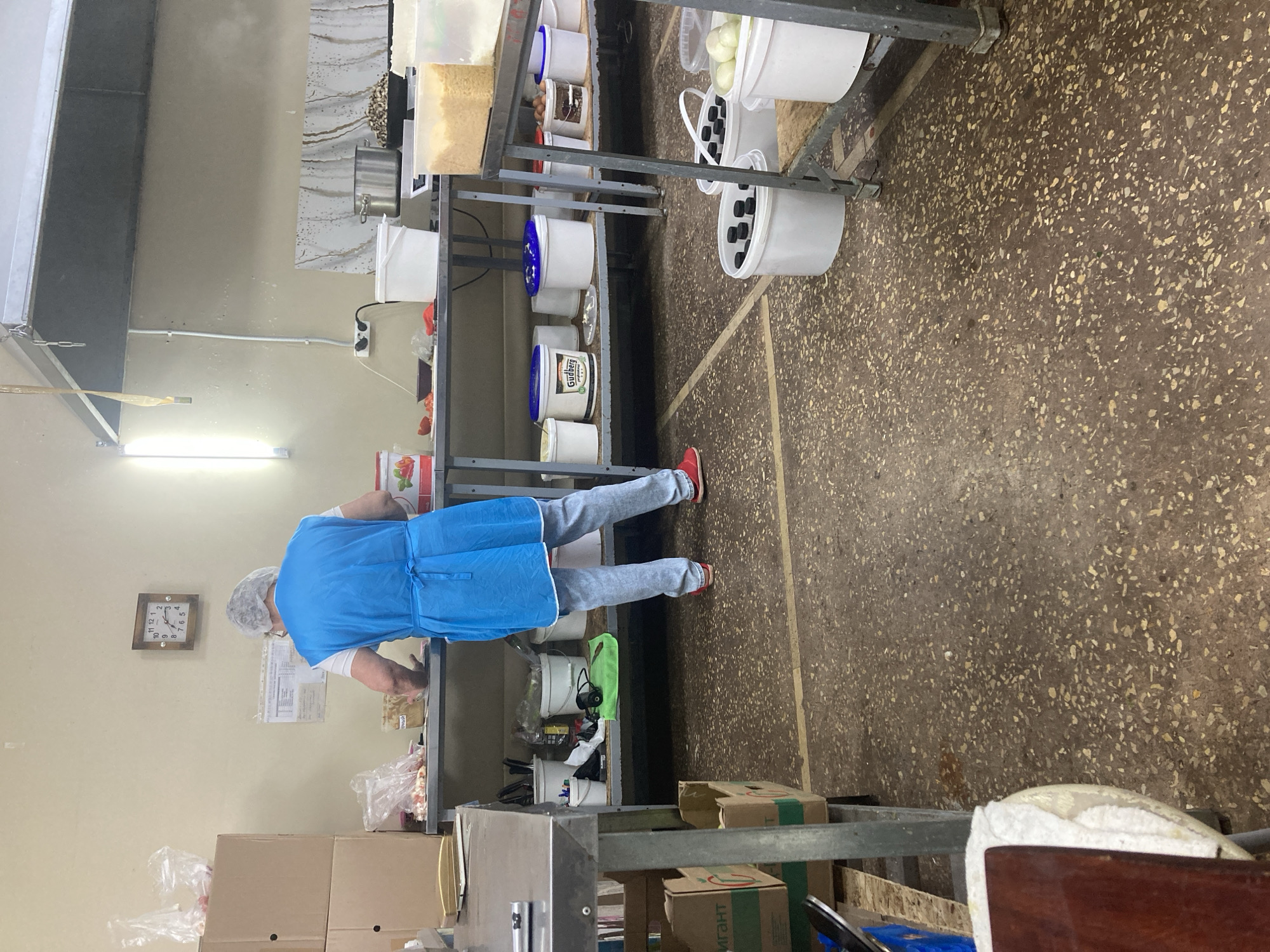 The image shows a person wearing a blue apron and hairnet working in a commercial kitchen, likely in Balakovo, Russia. The kitchen appears functional with stainless steel work surfaces and shelving units. Various food containers, buckets, and ingredients are visible on the shelves and floor. A clock on the wall suggests it is late afternoon. The floor is made of speckled concrete. There is no indication of the specific location in Balakovo, only that it is a kitchen environment.