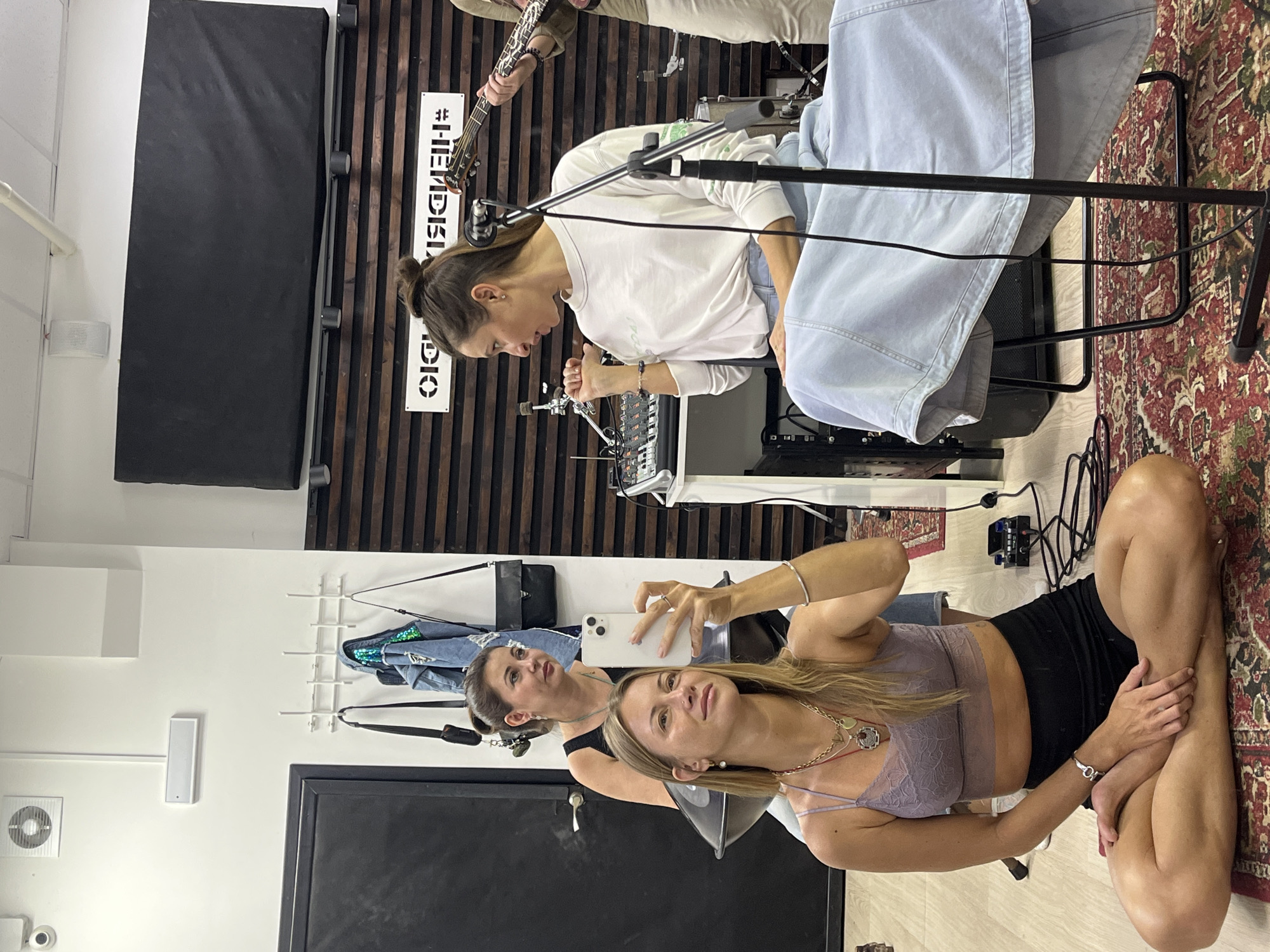 This media file captures a candid moment in what appears to be a music studio or rehearsal space in Moscow, Russia. Two women are the primary subjects. The woman in the foreground is seated on the floor, her legs crossed, looking up and to the side. She is wearing a light purple lace bralette and black shorts, with several necklaces and bracelets. A cell phone is held in her right hand, suggesting she may have just taken or is about to take a photo or video.
Behind her and slightly to the left, another woman is visible, also looking upwards and towards the left. She is wearing a white long-sleeved shirt. A guitar is visible being held by a person whose lower body is only partially in frame, suggesting a musical activity is underway.
The setting includes musical equipment, a mixing board, microphones, and what looks like acoustic paneling on the wall behind the musicians. The floor is covered with a patterned rug. The overall atmosphere appears relaxed and informal, possibly during a break in practice or recording. There is visible text on a sign on the wall which reads "#THEMBDEL". The lighting suggests it is daytime, though no specific weather cues are present.